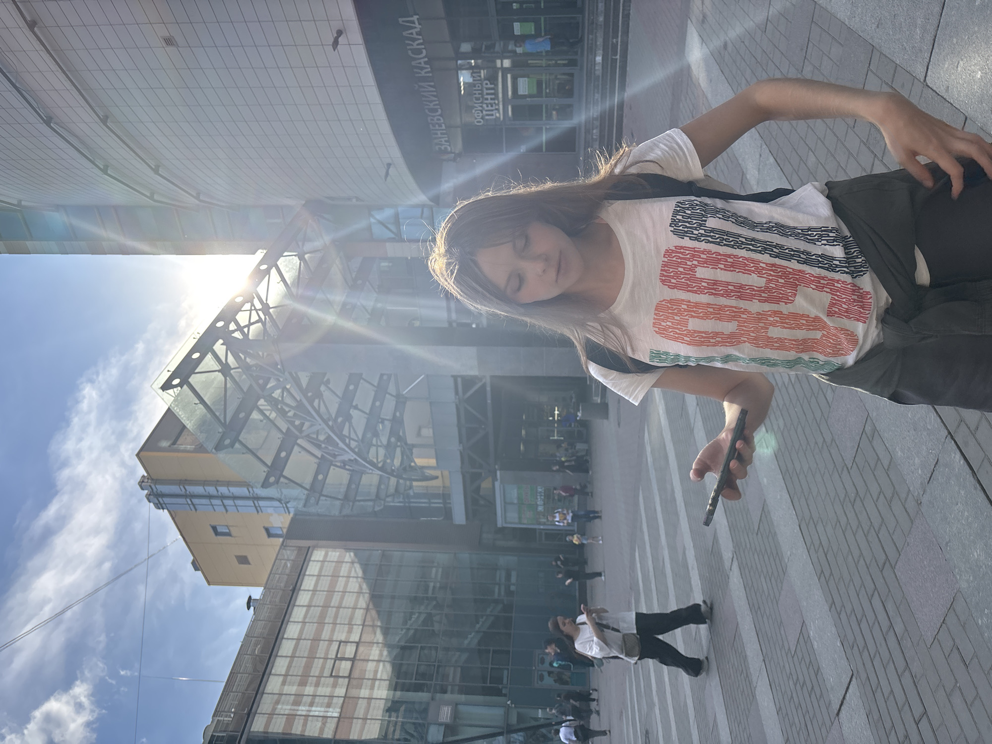 A young woman with light brown hair, wearing a white t-shirt with multicolored vertical text patterns, a dark backpack, and an olive-green jacket tied around her waist, stands on a paved urban sidewalk, looking down at a dark smartphone held in her right hand. Strong sunlight from the upper right illuminates the scene, producing visible lens flare. In the background, to the right, is the "Zanevsky Kaskad" (ЗАНЕВСКИЙ КАСКАД) business and shopping center in Saint Petersburg, Russia, identified by its curved, light-tiled facade and a prominent sign above an entrance which also reads "Office Center" (ОФИСНЫЙ ЦЕНТР). A contemporary glass and metal canopy structure extends from this building. To the left, another modern building with large reflective glass windows is visible. Several pedestrians are walking on the grey-patterned sidewalk in the mid-ground, which features distinct pedestrian crossing markings. The sky overhead is blue with scattered white clouds.