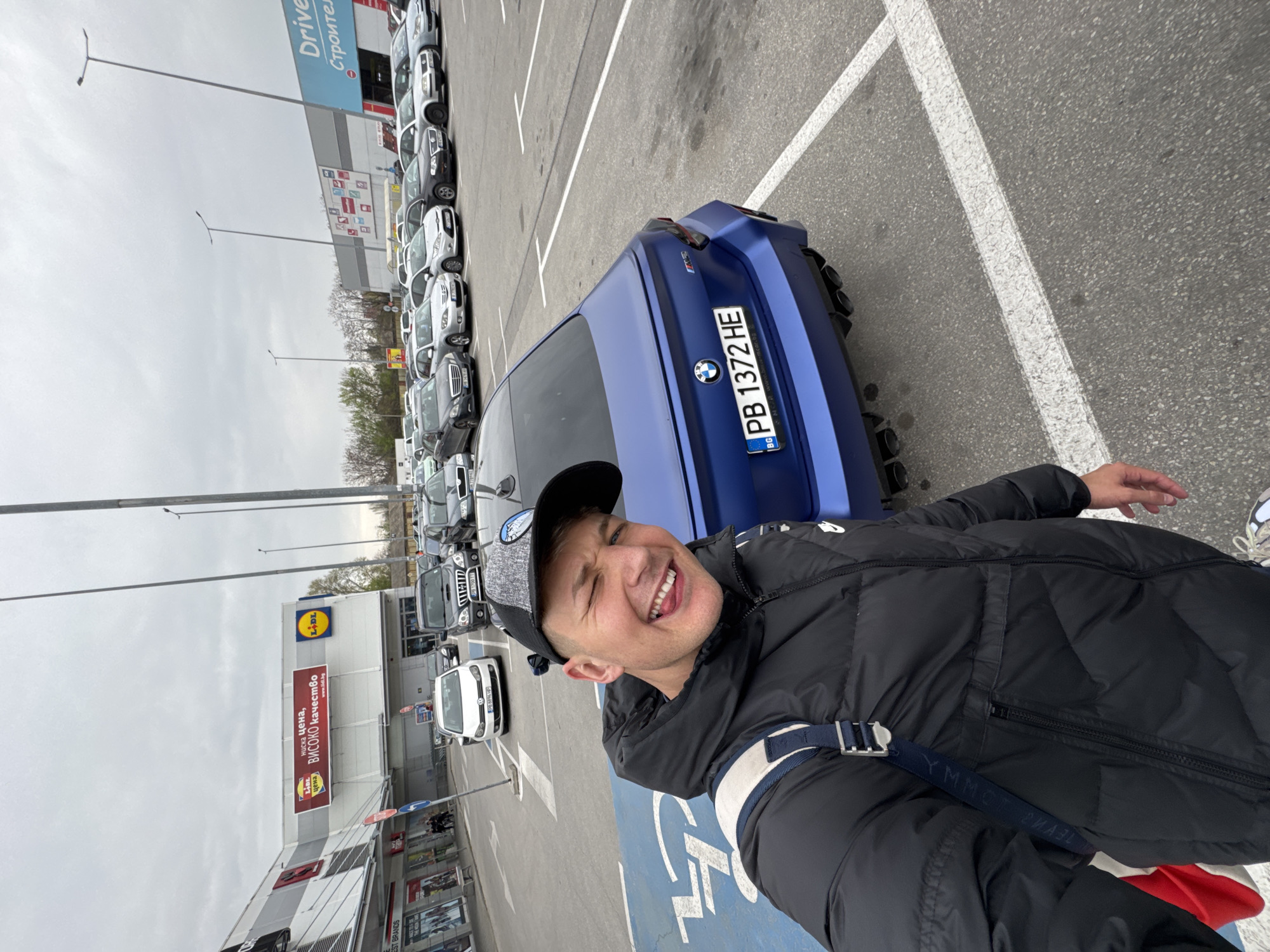 The image shows a man taking a selfie in a parking lot. He is wearing a black puffer jacket and a baseball cap. A blue BMW with license plate "PB 1372 HE" is parked behind him. In the background, there are rows of parked cars, a Lidl store, and a "Drive Строител" store. Based on the license plate, this image was likely taken in Plovdiv, Bulgaria.