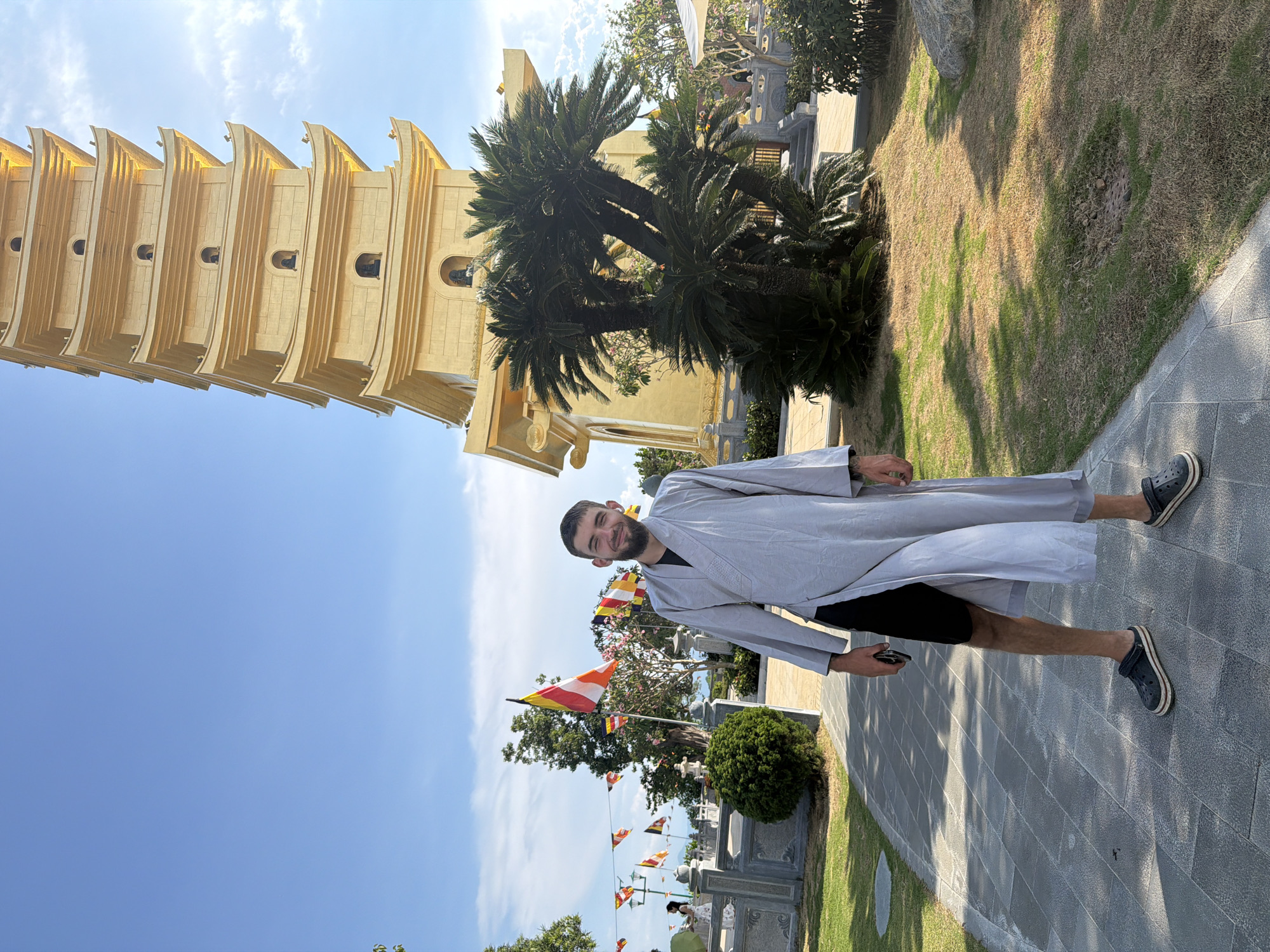 A man in a light gray robe stands on a paved path adjacent to a grassy area. To the left is a tall, golden pagoda. The sky is blue with some clouds. Buddhist flags are strung in the background. He is wearing dark shorts and dark shoes. The location appears to be in Nam Nha Trang Ward, Vietnam.