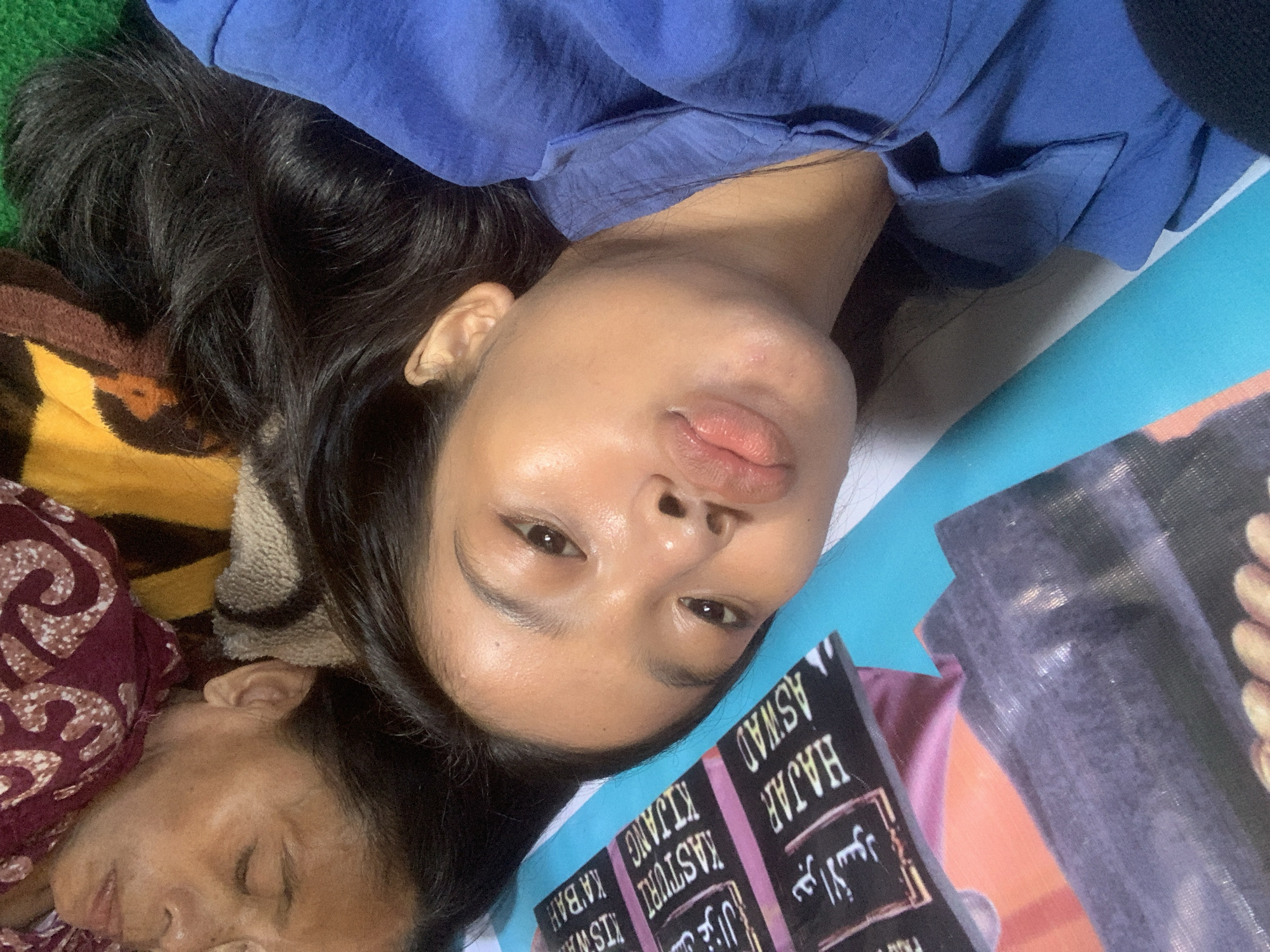 An intimate, tilted close-up shot captures two individuals resting indoors, likely in Sungai Meriam, Indonesia. The primary subject, a young woman, lies with her head turned, looking directly at the camera. She has long, dark hair, luminous skin, and full lips, wearing a blue top. Natural light highlights her face, creating a reflective sheen. Her expression is neutral to slightly pouting.
To her lower left, an older woman is visible, sleeping with her eyes closed. Her face is partially obscured by a maroon patterned fabric, suggesting she is covered by a blanket or sarong. The younger woman's head rests against a patterned fabric in shades of brown and yellow.
To the right of the younger woman, a dark-colored object, possibly a book or package, is prominently displayed. Visible text on this object includes "HAJAR ASWAD," "KASTURI," "KIJANG," "KISWAH," and "KA'BAH," written in both Latin and Arabic script, indicating items related to Islamic culture or pilgrimage. The background consists of a light blue surface and a hint of green fabric. The overall scene conveys a relaxed, casual moment, possibly a selfie or close-up photo taken during rest.