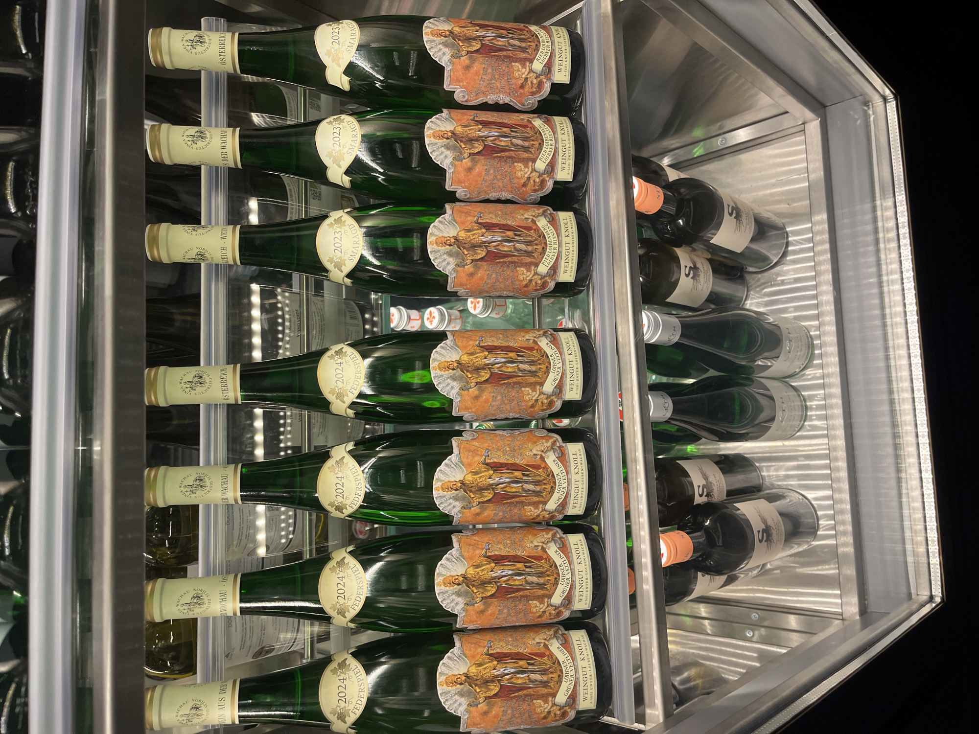The image presents a detailed view of the interior of a modern wine cooler or display case, well-lit and meticulously organized. The setting appears to be an indoor space, possibly in Sofia, Bulgaria, showcasing a collection of wines.
The primary subjects are numerous wine bottles, predominantly green-tinted, indicative of white wines, and some darker bottles, likely red wines. The green bottles are neatly stacked horizontally on clear, transparent shelves, while the darker bottles are arranged on shelves to the right. The cooler itself features a sleek, metallic (likely stainless steel) frame, with vertical LED lighting strips visible on the right side, illuminating the contents and creating reflections on the glass and polished surfaces.
A prominent feature of the green bottles is their elaborate main labels, which depict a richly adorned historical figure, possibly a king or emperor, surrounded by cherubs. The text "WEINGUT KNOLL" is clearly visible on these labels, identifying the Austrian winery. Further details on these labels include specific wine types such as "LOIBNER GRÜNER VELTLINER" and "LOIBNER RIESLING," along with "RIED LOIBENBERG" and the winery's location, "A-3601 UNTER-LOIREN WACHAU." Smaller neck labels on these bottles indicate vintages and quality classifications, such as "2023 SMARAGD" and "2024 FEDERSPIEL," with "WACHAU" and "ÖSTERREICH WEIN" also visible, confirming their Austrian origin. The darker bottles on the right have less discernible labels, though a white label with a stylized 'S' is somewhat visible.
No people are present, and the image solely focuses on the presentation and storage of the wine bottles within the sophisticated cooler, emphasizing organization and the quality of the displayed products. The internal lighting highlights the bottles and their intricate labels.