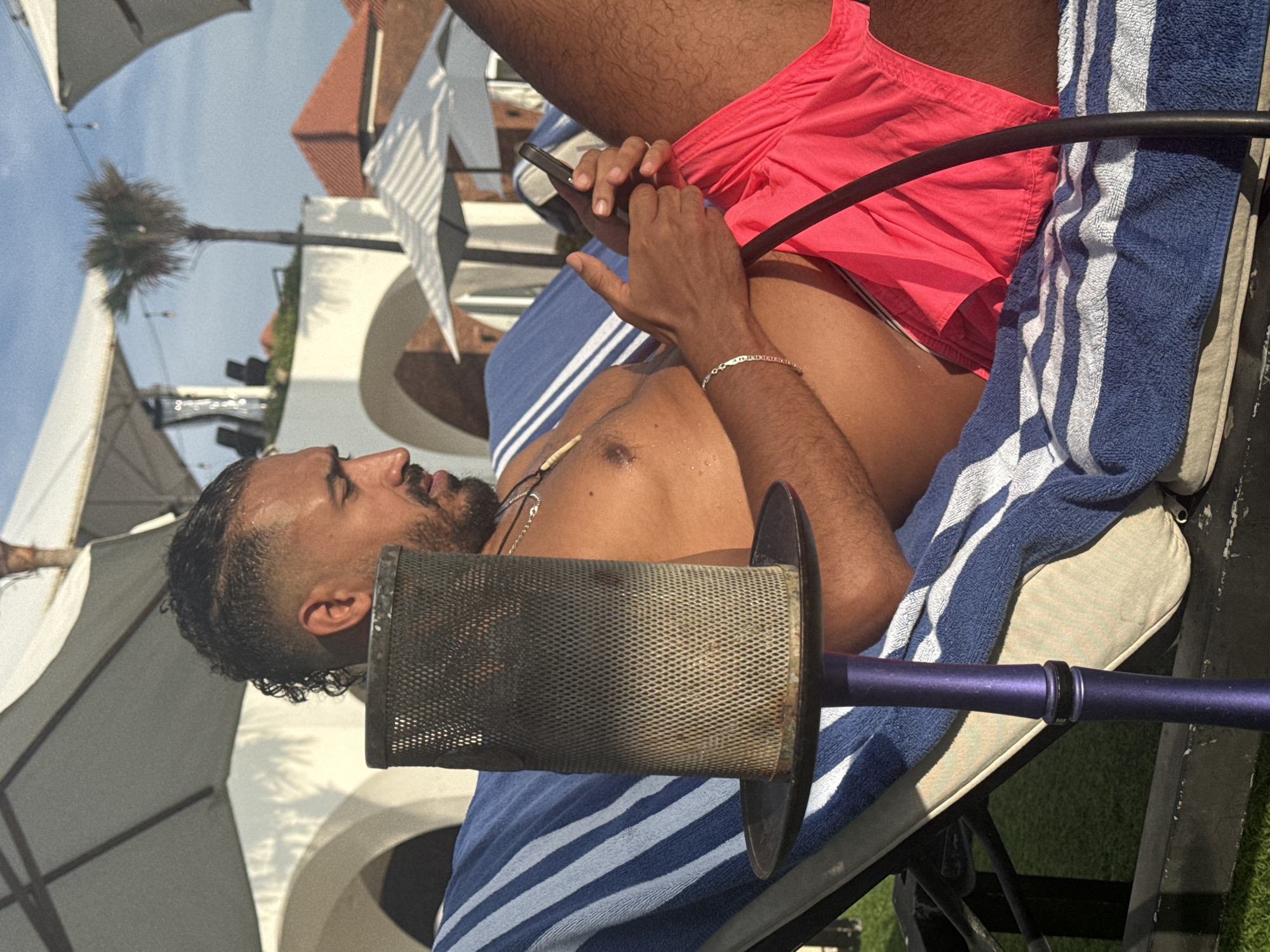 A man reclines on a striped blue and white towel draped over a light-colored lounge chair, holding a mobile phone in his hands. He is wearing pink shorts and a silver bracelet, and a hookah stem is positioned near his face. In the background, palm trees, white buildings with arched openings, and patio umbrellas suggest a resort or poolside setting in North Kuta, Indonesia.
