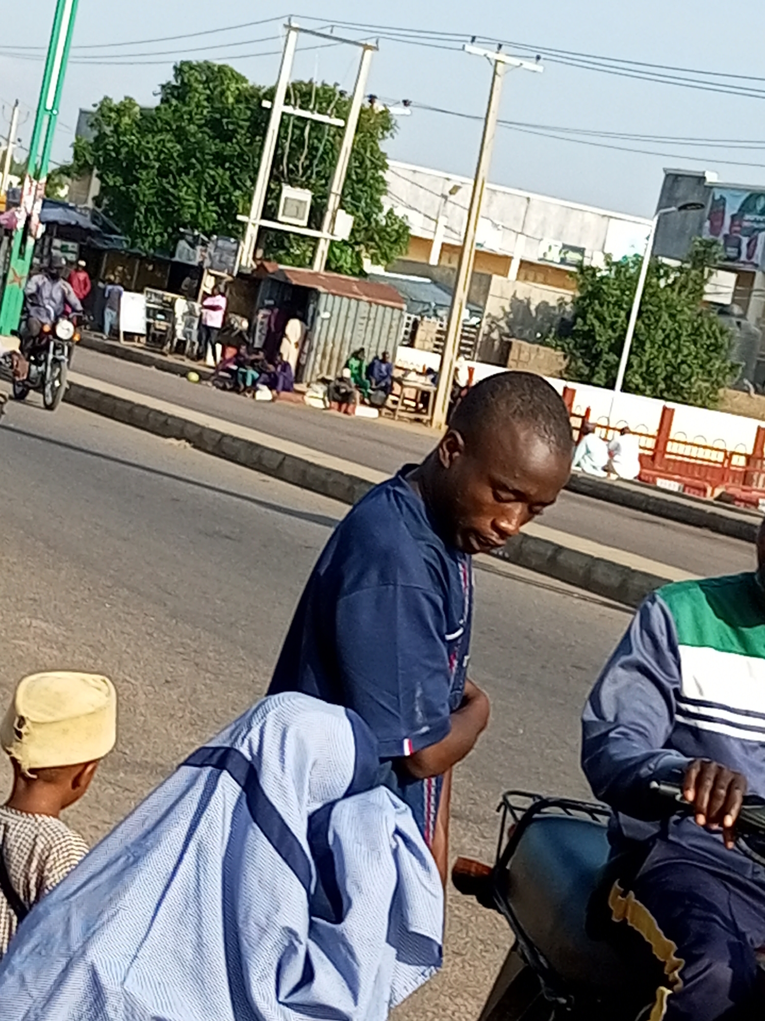 This is a medium-distance, slightly blurry, eye-level shot of a street scene in Tumfure, Nigeria. In the foreground, a man in a dark blue t-shirt is looking down, seemingly preoccupied. To his left, a young child wearing a light-colored checkered outfit and a yellow cap is visible from the back. Partially obscuring the child and the man is a person in a light blue, striped garment. To the right, a person is seated on a motorcycle, wearing a jacket with green and white stripes on the sleeves and dark pants. In the middle ground, a motorcycle is moving towards the left of the frame. The background features a bustling street with various small shops, people in traditional clothing, and trees. Power lines and utility poles are prominent in the upper portion of the image. The overall impression is of a typical, busy street environment.