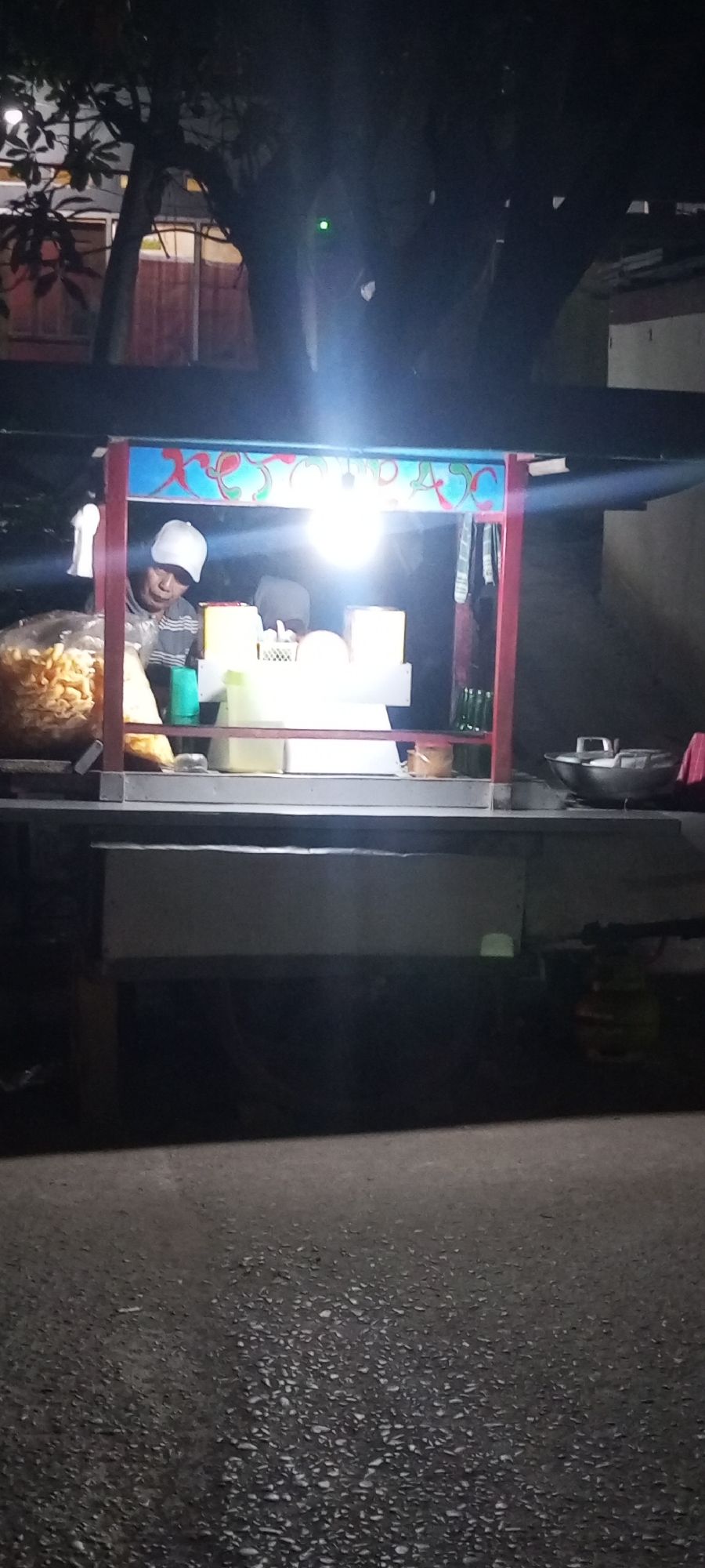 A man wearing a white cap and a striped shirt is standing behind a food cart. The food cart is lit by a bright overhead lamp and has colorful decorations. There are bags of food items on the cart, and various containers are visible on its surface. To the right of the cart, a large metal wok and a red bag are present. The scene appears to be outdoors at night, with trees and a building visible in the background. The ground in front of the cart is paved with asphalt. The food cart is likely situated on a street or sidewalk in Setu, Indonesia, possibly near a residential area or a marketplace.