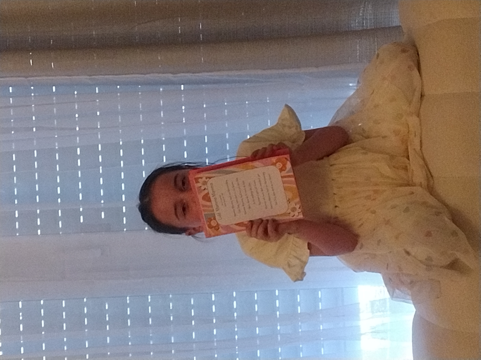 The image shows a young girl holding a book. She is wearing a light-colored dress with a floral pattern. The girl is positioned in front of a window with white blinds. The scene is brightly lit, suggesting it is daytime. The girl appears to be reading or showing the book to someone off-camera. The book has a pink and yellow cover with a floral design. Visible text on the book reads "Buona Mamma". The overall atmosphere is casual and domestic, possibly within a home in Crauthem, Luxembourg.