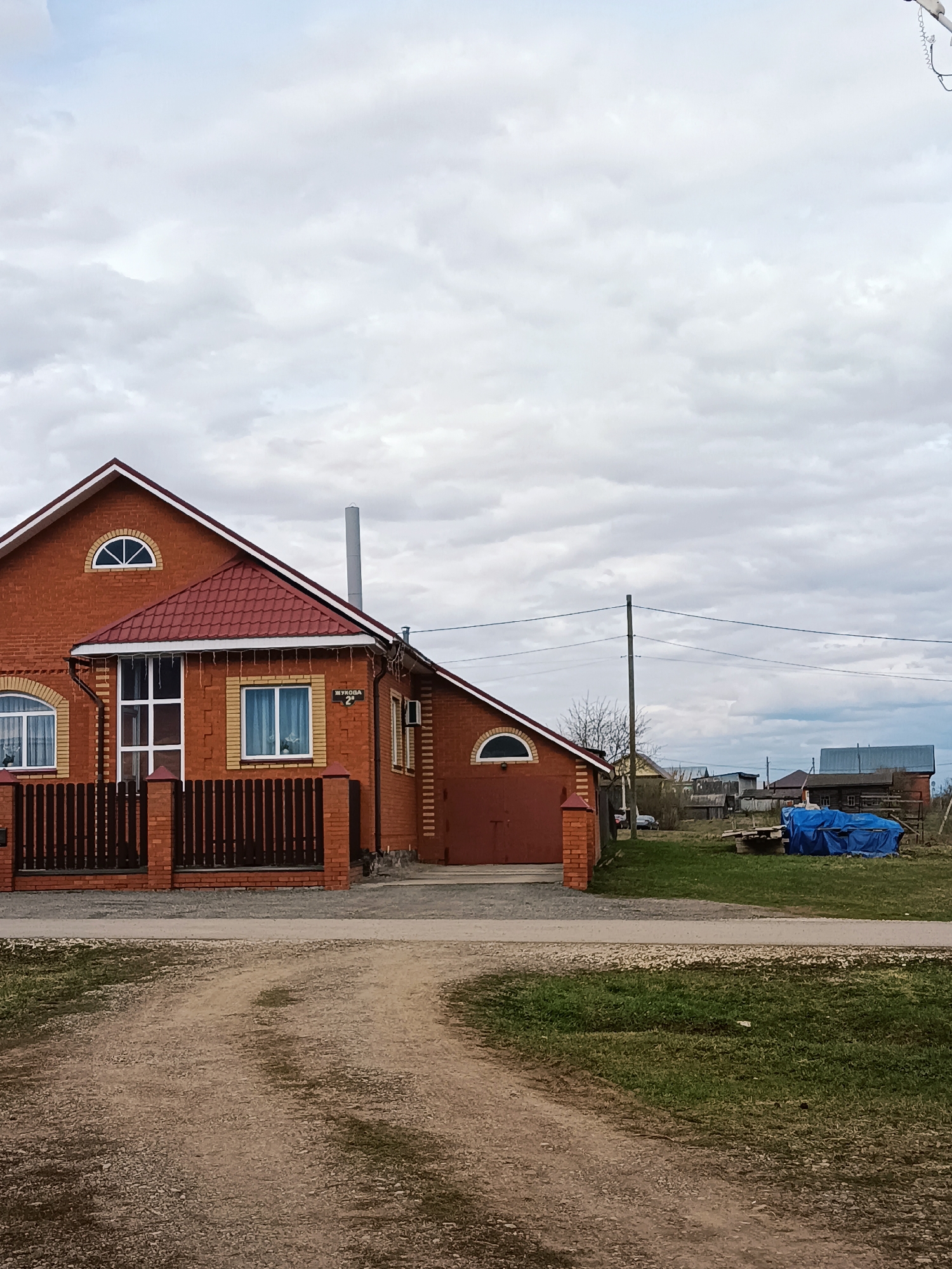 The image displays a daytime view of a residential property in a rural or semi-rural setting, likely in Barda, Russia. The sky is heavily overcast with thick, light grey clouds, indicating no direct sunlight.

The main subject is a substantial two-story house constructed from reddish-brown bricks, accented with lighter-colored brickwork around windows and corners. It features a prominent red tiled roof, a tall grey metal chimney, and several windows, including a distinctive semicircular arched window on the gable end. The house has a garage with dark red double doors on its right side. A dark brown wooden fence with brick posts encloses the front of the property.

In the foreground, a light-colored dirt path or driveway curves from the bottom left, leading towards the house. To the right of this path is a strip of green grass. A utility pole with power lines stands to the right, extending across the mid-ground. Further back, on a grassy area, a blue tarp covers a pile of items. Several other distant, simpler buildings and sparse trees are visible, suggesting a village or small town environment.

A white sign with black Cyrillic text is visible on the brick wall of the house, to the right of a large window. The text reads "ЖУКОВА" (Zhukova) followed by the number "2", which likely indicates a street name and house number. No people are visible in the scene.