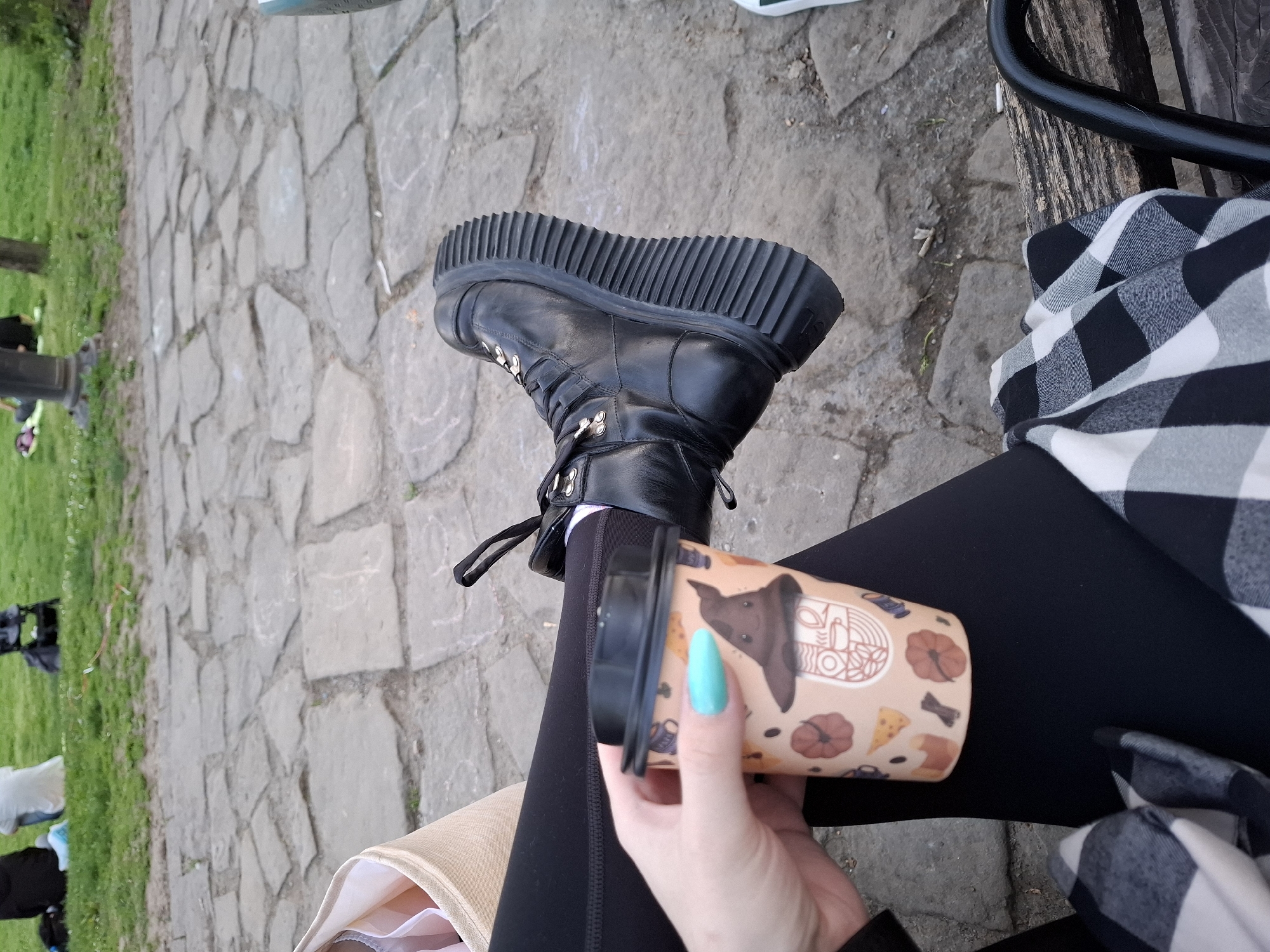 A person is sitting on a bench, holding a coffee cup with a Halloween-themed design, resting their leg with a black platform boot across the other leg. The person is wearing black leggings and a black and white checkered shirt. The bench appears to be in a park setting, with a stone path visible in the background. The photograph was taken in Novi Sad, Serbia.