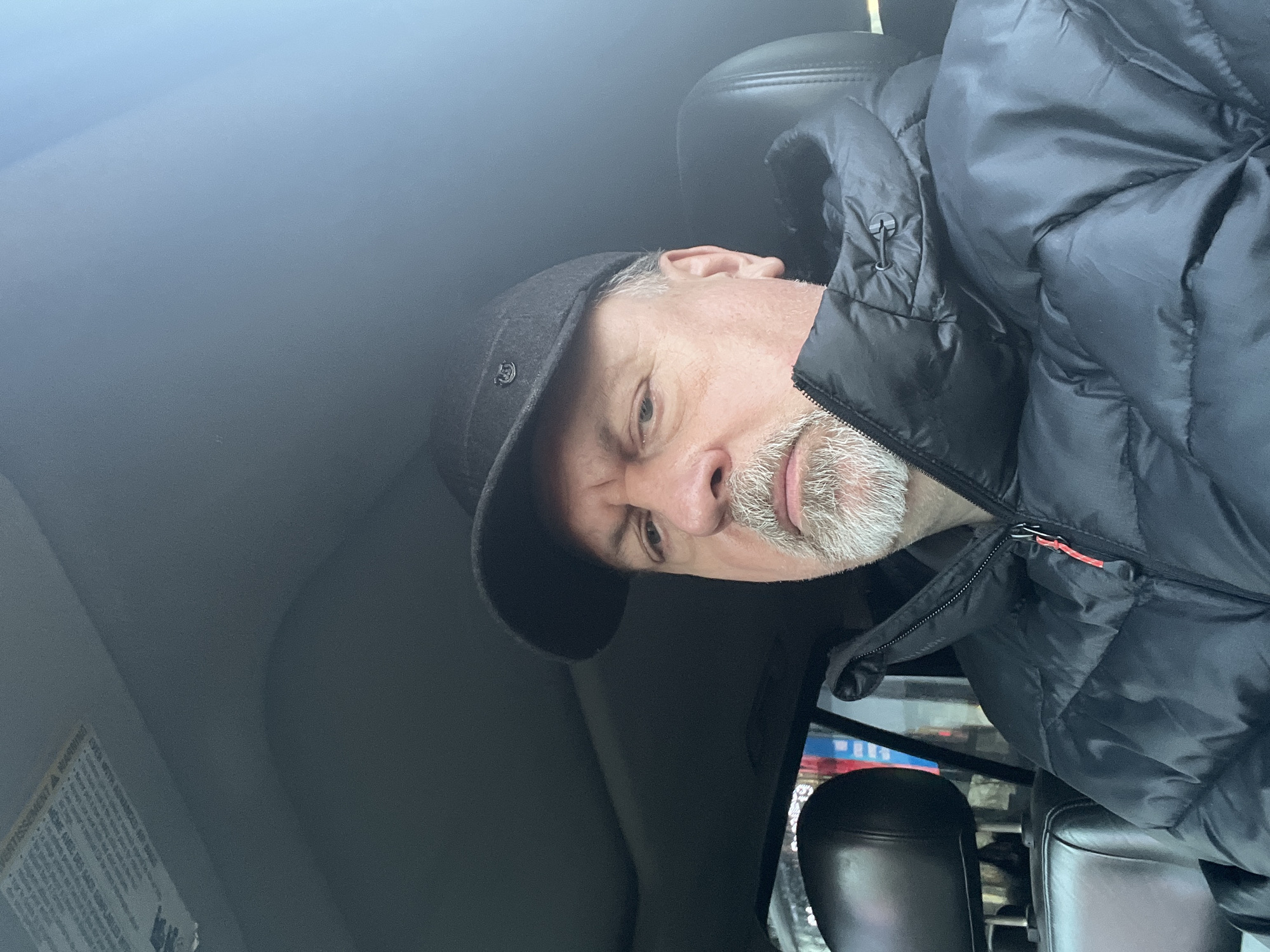 A middle-aged man with a gray beard and wearing a dark baseball cap and a black puffer jacket is seated in a vehicle. The vehicle's interior is visible, including a headrest and part of the dashboard. The man is looking towards the camera. The location of the photograph is not apparent, and there is no information to determine the city of Podolsk, Russia.