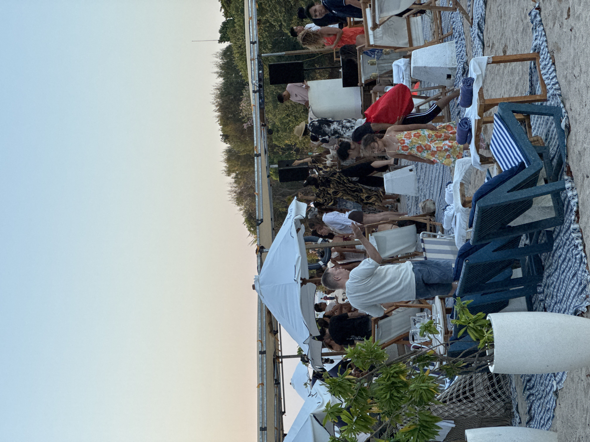 The image depicts a beach party scene at sunset in Kuta Selatan, Indonesia. The beach is set up with lounge chairs, white umbrellas, and decorative rugs on the sand. In the background, a DJ booth with speakers is visible, and people are gathered around, some dancing and socializing. The sky is a gradient of pale blue to orange, indicating the time of day. The environment is a typical beach resort atmosphere with lush green trees bordering the sand.