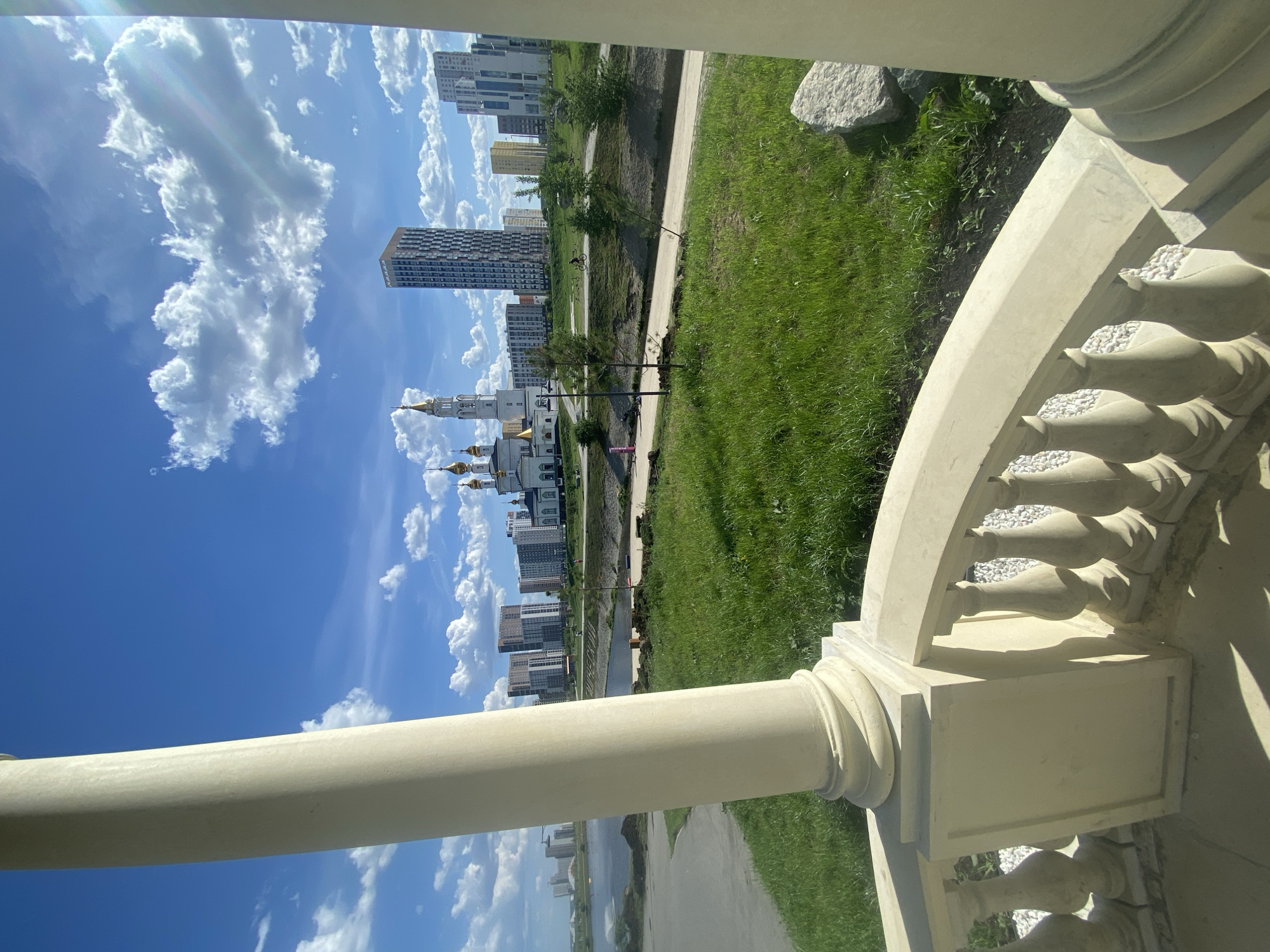 The image shows a view from within a balustrade, likely part of a building or structure. Through the balustrade, there's a view of Yekaterinburg, Russia. In the middle ground, there is an Orthodox Church, followed by apartment buildings. There is a river, grass, and walkway visible. There are clouds in the blue sky. The foreground consists of the balustrade itself, made of stone or concrete, with classical design elements. There are no people visible in the photo.