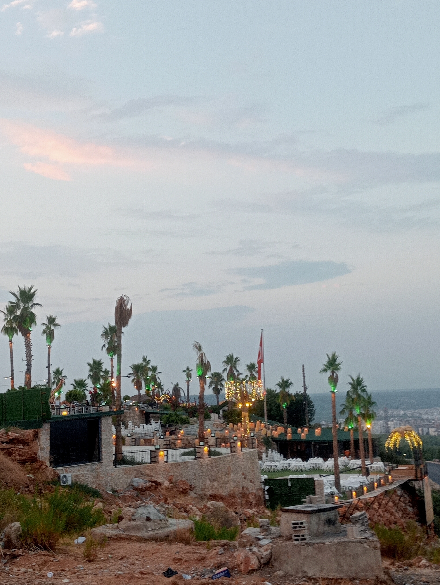The image captures an elevated view of a resort or restaurant establishment situated on a hillside overlooking a city, likely Kepez, Turkey. The scene is set during dusk, with a soft, diffused light from the sky painting pastel hues across the clouds. The establishment features multiple levels with outdoor seating areas adorned with numerous lights. Palm trees are a prominent feature, some illuminated with green lights. Several buildings, likely part of the resort, are visible, including a larger, dark-roofed structure in the midground and a smaller building with an open entrance in the foreground. There are no people visible in the frame. The foreground is characterized by a rocky, uneven terrain with sparse vegetation and some scattered debris. The background reveals a distant cityscape and the calm expanse of the sea. A Turkish flag is visible on a flagpole, adding a nationalistic element to the scene.