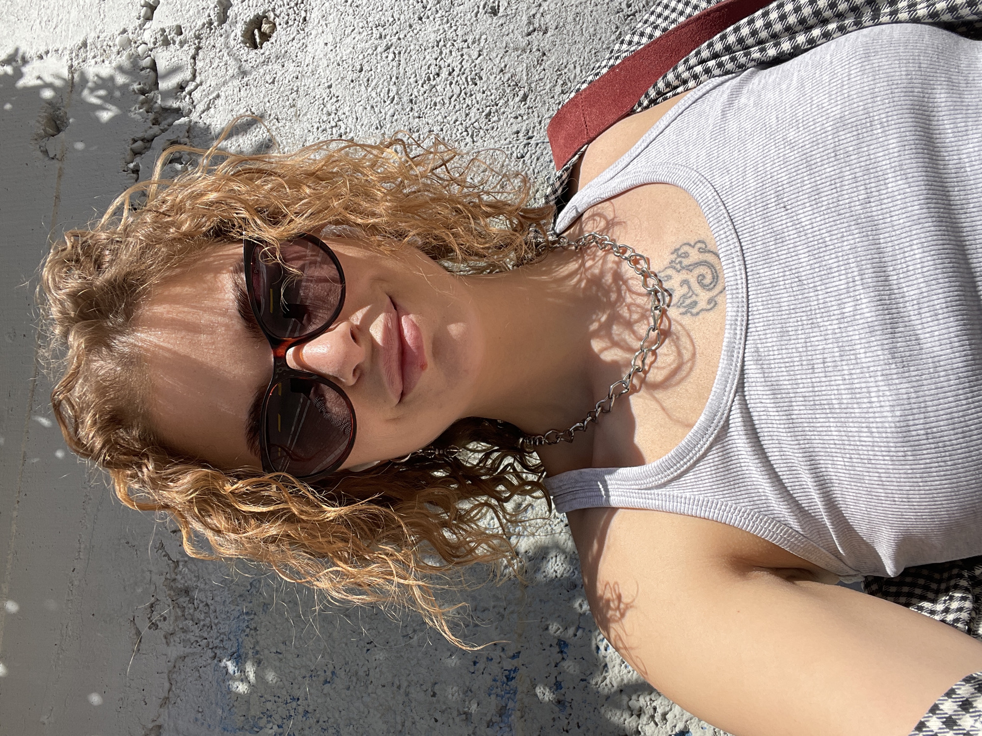 A young woman lies on a concrete surface outdoors, wearing a light gray tank top, a silver chain necklace, and sunglasses. A portion of her checkered jacket is visible. She has curly blonde hair. A tattoo is visible on her chest near her shoulder. The location appears to be outdoors in the city of Rafailovići, Montenegro, based on the concrete surface, the shadows cast by trees, and likely architectural features.