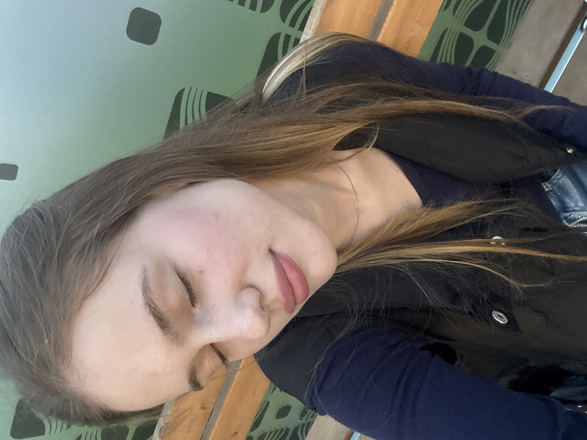 A young woman is captured in a close-up, slightly angled selfie. Her eyes are closed, and her head is tilted down, revealing a soft smile. She has long, brown hair with lighter streaks, which falls around her face and shoulders. She is wearing a dark blue long-sleeved shirt and a black vest. The background is a blurred green surface with a repeating abstract pattern of dark shapes, possibly a wall or screen. To the left and right of the subject, sections of a wooden bench are visible, suggesting she might be seated outdoors or in a waiting area. The overall lighting is soft, highlighting her skin and makeup, which includes pink blush and lip color. There is no indication of the city of Arfa, Russia, in the image.