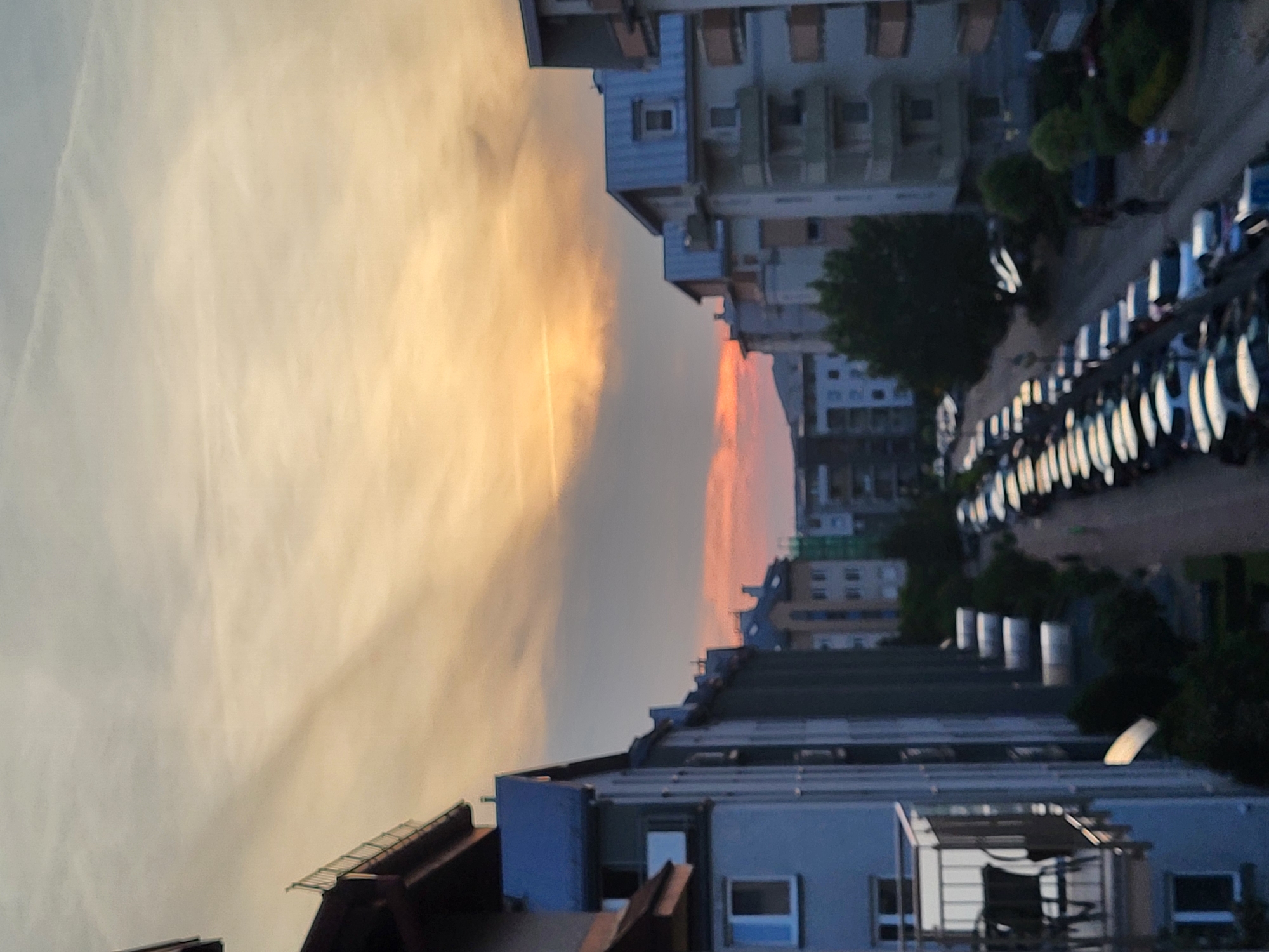 The image captures an urban landscape at sunset, viewed from an elevated perspective. The scene is dominated by a dramatic sky, which displays a gradient of colors from light blue and pale yellow in the upper left, transitioning to vibrant orange, pink, and reddish hues at the horizon on the left. A distinct band of dark grey cloud stretches horizontally across the mid-ground, separating different atmospheric layers.
Buildings of various heights and architectural styles line the right side and bottom of the frame, appearing as multi-story structures with visible windows and balconies. Many of these buildings are lit with the soft, warm light of the setting sun. Along the bottom right, a street or parking area is visible, densely populated with parked vehicles, possibly cars or motorcycles, whose surfaces reflect the diminishing light. Interspersed among the buildings and along the street are green trees and bushes, adding touches of nature to the urban environment. The overall impression is one of a peaceful evening setting in a residential or mixed-use urban area. No specific human activity is depicted, and no visible text is present.