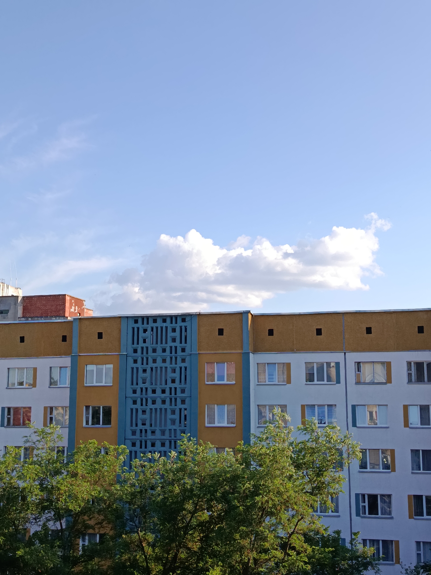 The image shows a residential apartment building in Svietlahorsk, Belarus. The building has a light grey facade with bands of yellow and blue. The windows are arranged in rows, and some have blue shutters. A decorative blue panel with a geometric pattern is located in the center of the building. Trees with green foliage are in the foreground, obscuring the lower portion of the building. The sky above is blue with scattered white clouds. The overall impression is that of a typical urban residential scene in a post-Soviet city.