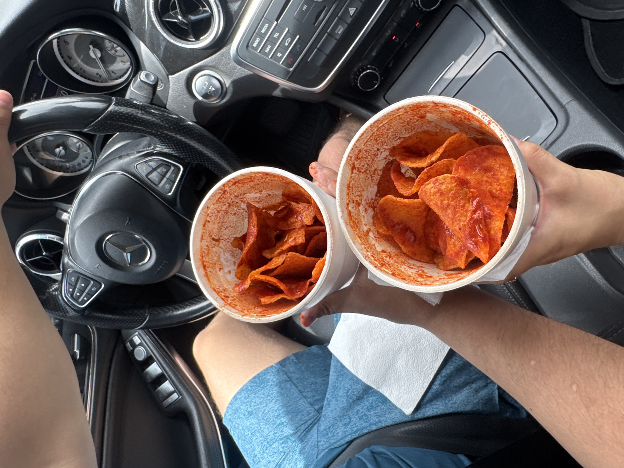 The image shows the interior of a Mercedes-Benz vehicle. A person, presumably the driver, is holding the steering wheel with their left hand. In the passenger area, another person is holding two cups of Tostitos Preparados. The cups are white and filled with the chips covered in a red sauce. A white napkin is visible beneath the cups. The image was taken in Puebla, Mexico.