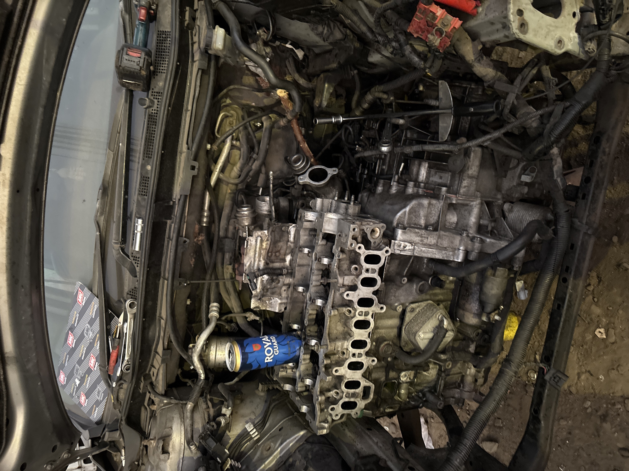 The image shows a disassembled car engine bay. The engine is partially disassembled with various components visible, including the engine block, cylinder head, intake manifold, and numerous hoses and wires. A blue Royal Guard can is positioned within the engine compartment. Various tools are scattered around the engine bay, including wrenches, sockets, and a power drill. The vehicle's windshield and dashboard are visible on the left side. The environment appears to be a garage or workshop, with a cluttered and disorganized workspace. There are no people visible in the image. The location in the city of Arica, Chile, is not determinable from the given image.