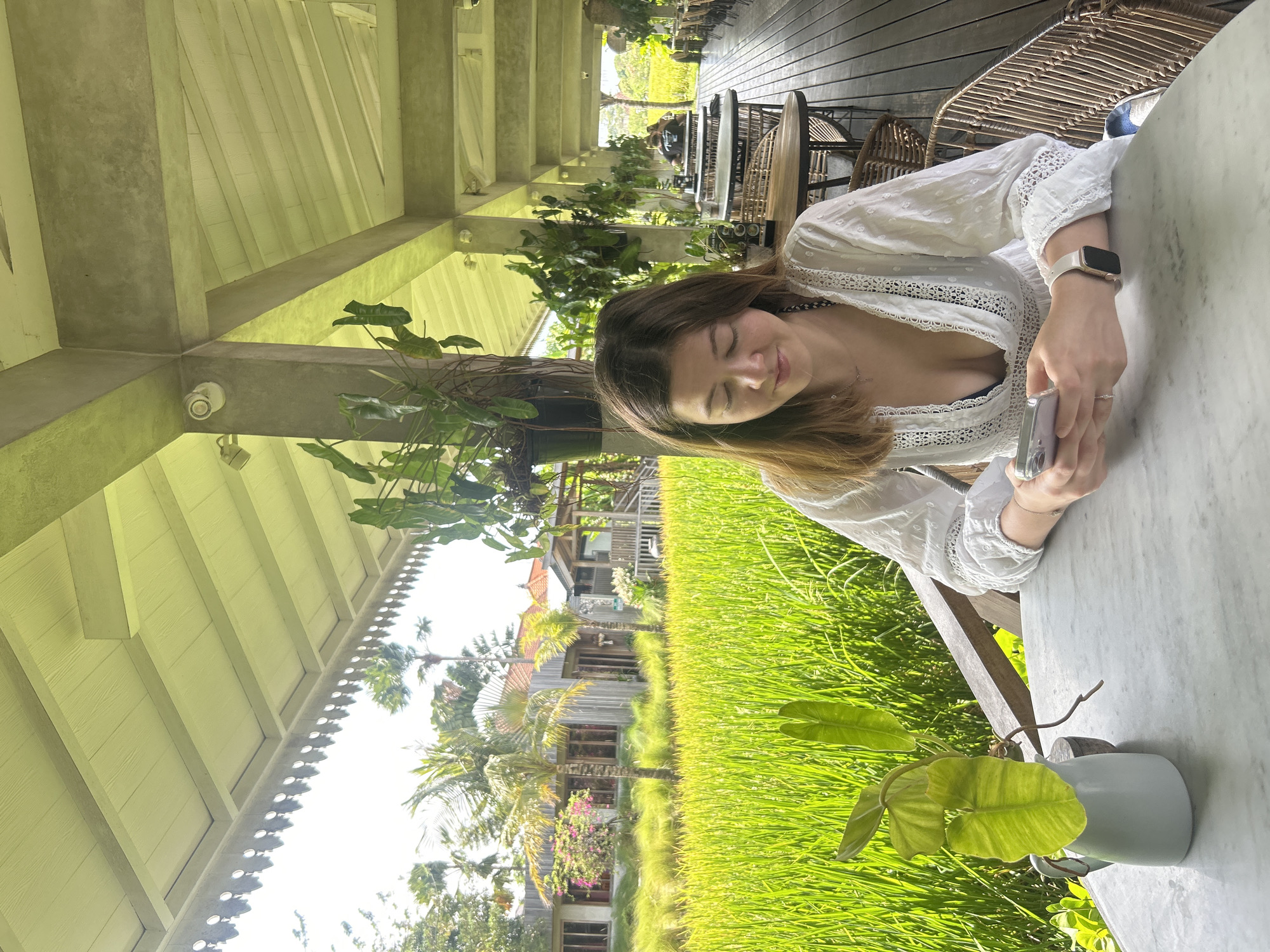 A young Caucasian woman with brown hair is seated at a marble-topped table on a covered patio overlooking a bright green rice field. She is wearing a white eyelet dress with a black bikini top visible underneath. She is looking down at her smartphone, which she holds in her hands. A small plant in a white ceramic vase sits on the table to her left. The patio has a wooden floor and is furnished with rattan chairs and tables. In the background, beyond the rice field, are traditional Indonesian buildings with red-tiled roofs, surrounded by palm trees and lush vegetation. The sky is bright and sunny. The woman is wearing a smartwatch on her left wrist. The general location is a restaurant or cafe in North Kuta, Indonesia, with a view of rice paddies.