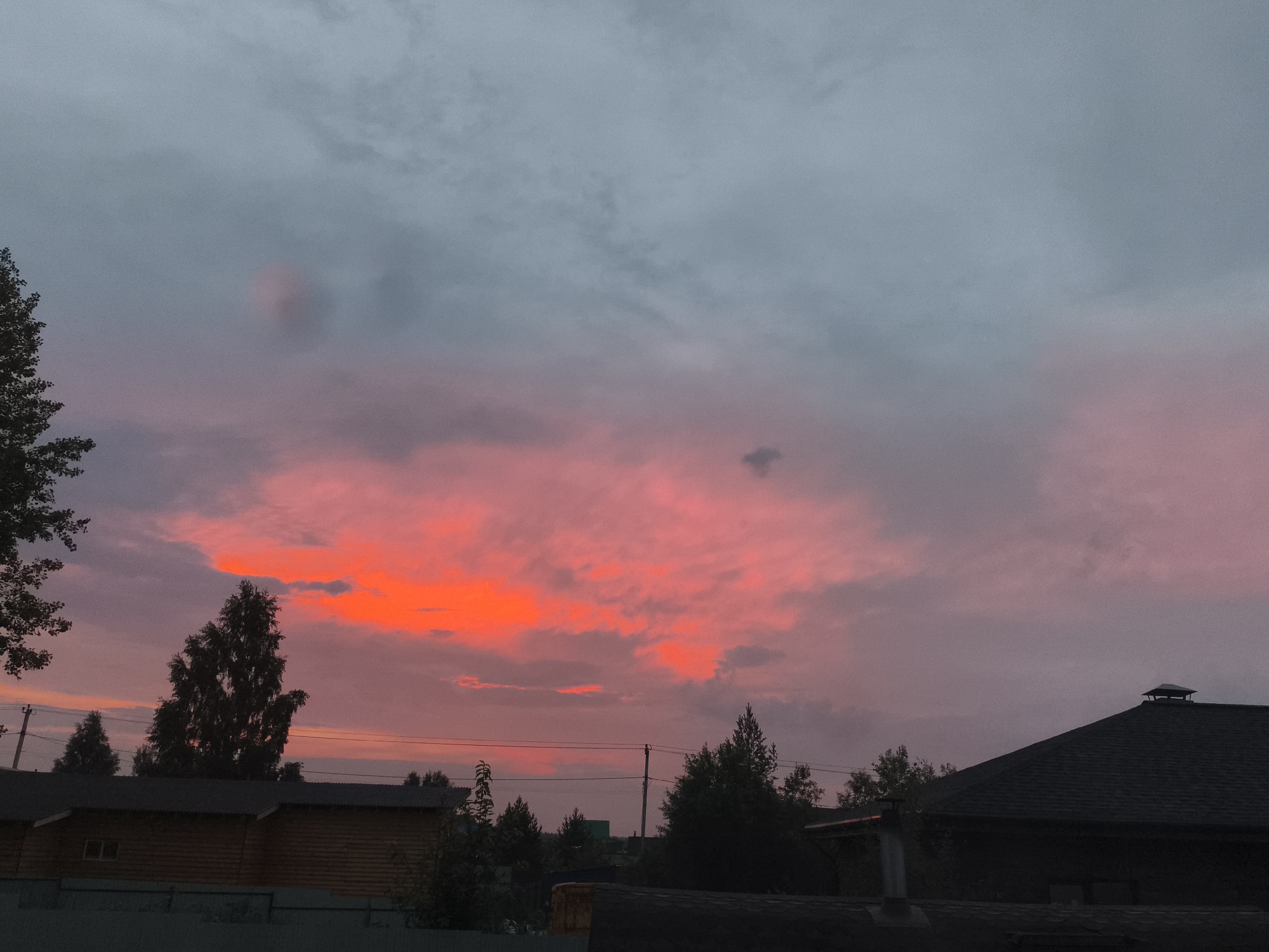 The image captures a vibrant sunset over a rural landscape in Русский Вожой, Russia. The sky is dominated by dramatic clouds, with a large portion glowing with intense orange and red hues, suggesting the sun is low on the horizon. The rest of the sky is a gradient of pink and muted gray, indicating the transition from day to night.
In the foreground, silhouetted against the bright sky, are the roofs of several buildings, likely houses, with dark, textured surfaces. One building on the left is made of horizontal wooden logs, with a small window visible. Another building on the right has a dark, sloped roof with a small cap on its peak. Trees, dark and leafy, punctuate the scene, with a tall, prominent tree in the center left and several smaller ones scattered across the middle ground and background. Power lines stretch across the sky, adding to the rural feel of the setting.
The overall mood is serene and picturesque, characteristic of a twilight scene. The intense colors of the sunset evoke a sense of awe and tranquility. There is no visible text in the image. The time of day is clearly sunset or just after, with the weather appearing calm and clear, apart from the dramatic cloud formations.