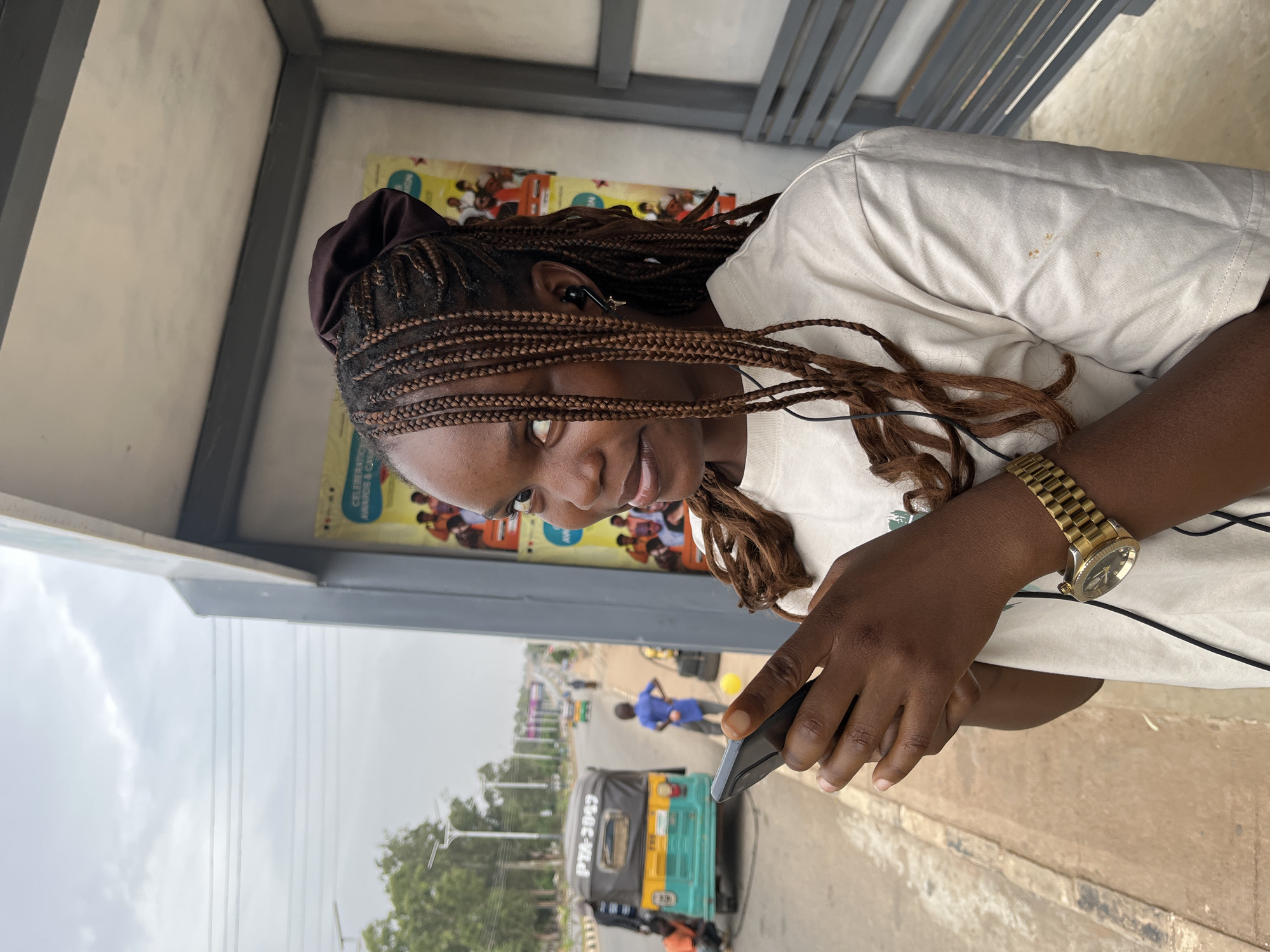 A young woman with braided hair sits at a bus stop in Bar Jirgi Summa, Nigeria. She is holding a smartphone in her left hand and wearing a gold wristwatch on her left wrist. She is wearing a light-colored t-shirt. An "okada" or auto-rickshaw is parked on the side of the road near the bus stop. In the background, there is a street scene with people and buildings.