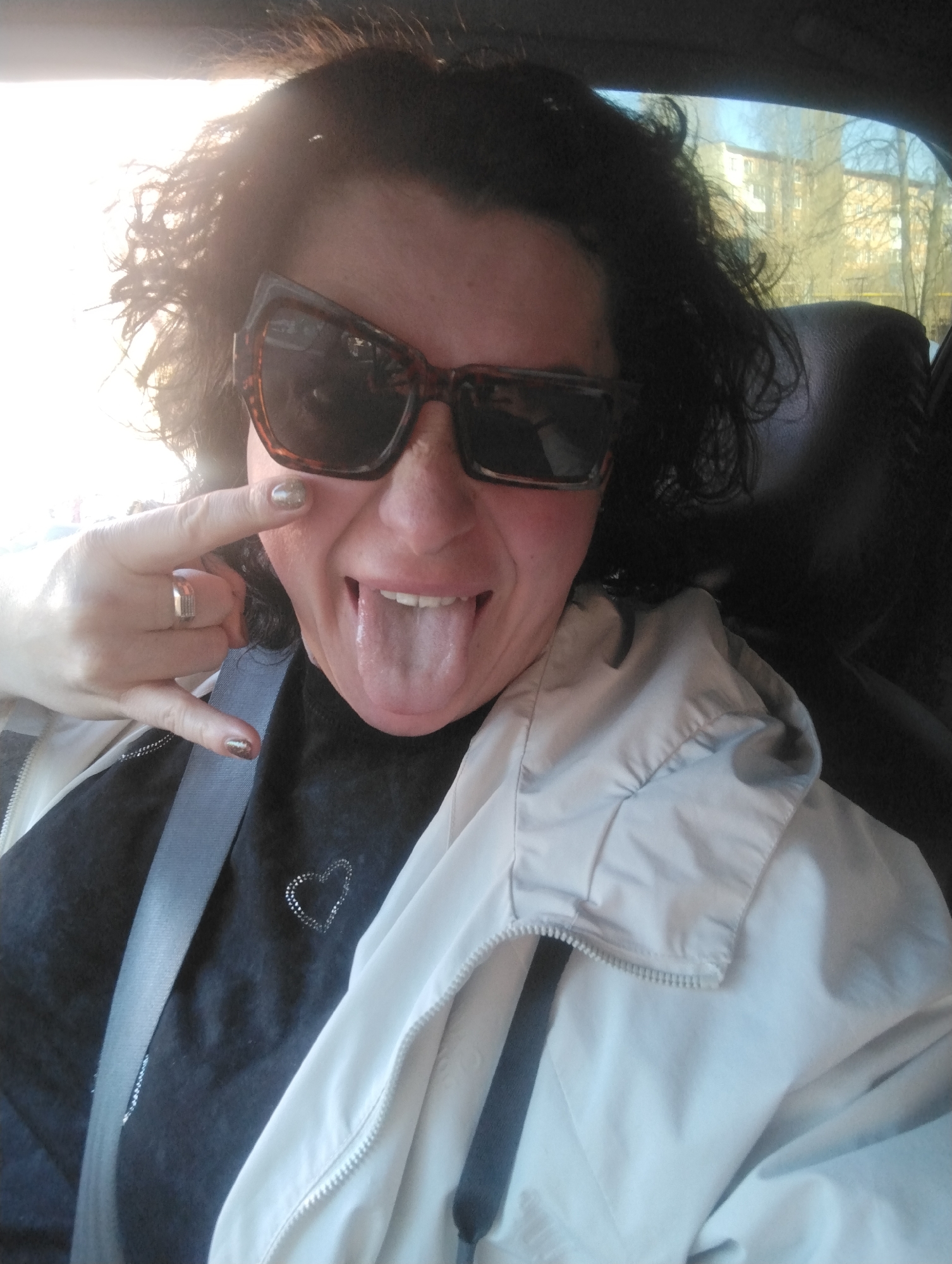 A woman with dark curly hair and oversized brown sunglasses is taking a selfie indoors, likely in a car. She is wearing a black top with a small heart design and a light beige jacket. She is wearing a seatbelt. She is sticking out her tongue and making a rock and roll hand gesture. A building is visible outside through the window, which, taken with the prompt about location, indicates the photo was taken in Votkinsk, Russia.