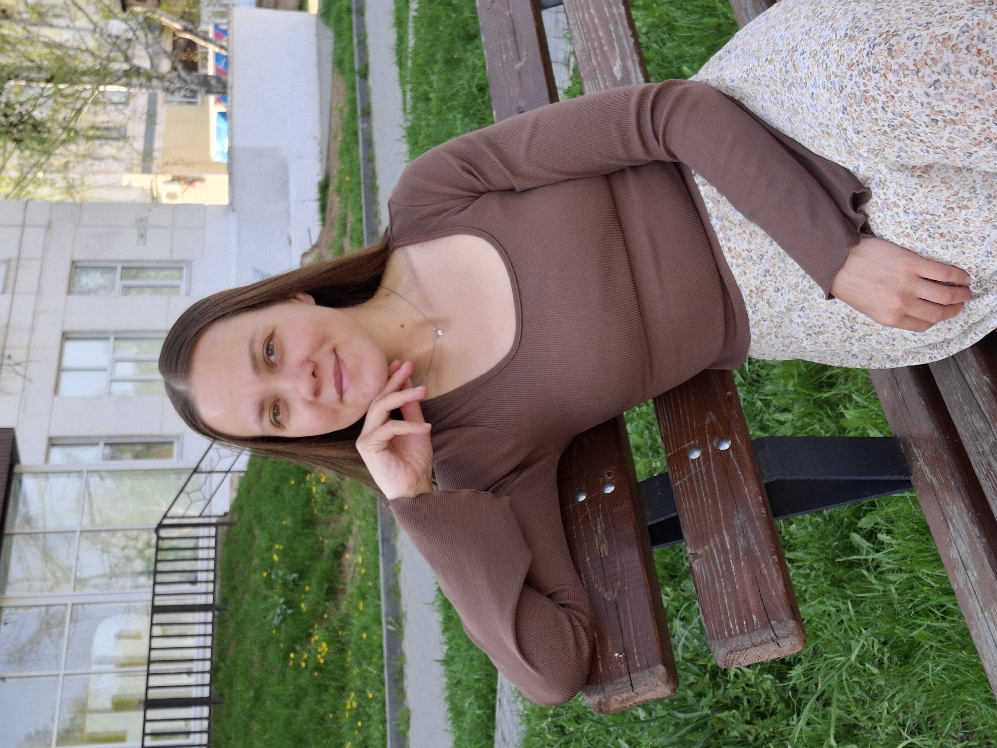 A woman is lying on a park bench in Chaikovsky, Russia. She has long brown hair and is wearing a brown long-sleeved shirt and a floral print skirt. She is resting her head on her hand and looking at the camera. The park bench is made of wood and has a metal support. The ground around the bench is covered in green grass and small yellow flowers. In the background, there is a light-colored building with windows.