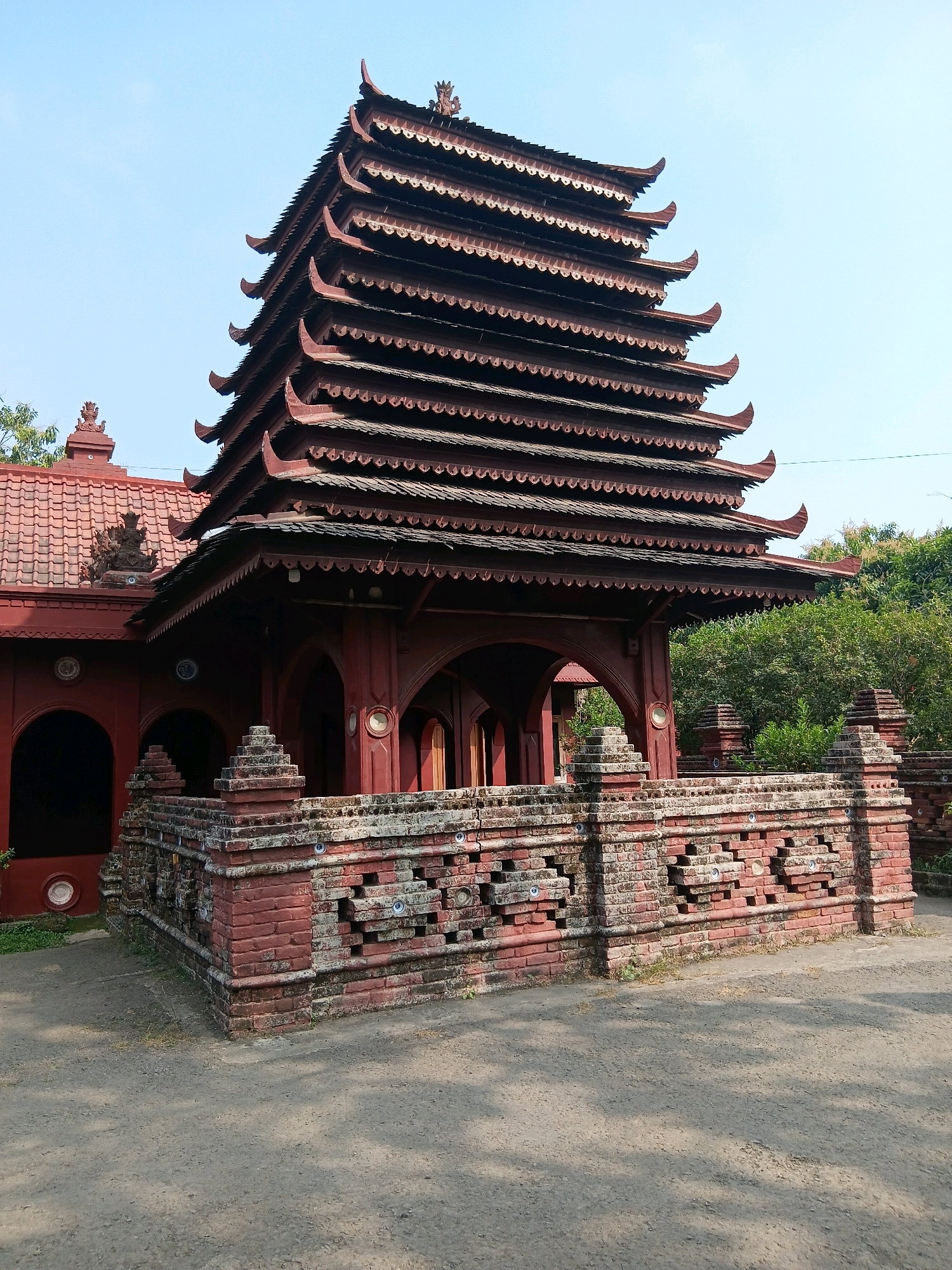 The image shows a multi-tiered, dark red pagoda-like structure with several overlapping roofs, characteristic of traditional Javanese architecture. The structure is made of wood and has ornate decorative elements on its edges. It is surrounded by a low, red brick wall with patterned openings and is situated in an outdoor environment with a clear blue sky and lush green trees in the background. The ground in front of the structure is a dirt path. The building appears to be a historical or cultural site, possibly a temple or a similar religious or ceremonial building. This structure is located at Sunan Gunung Jati tomb complex in Cirebon, Indonesia.