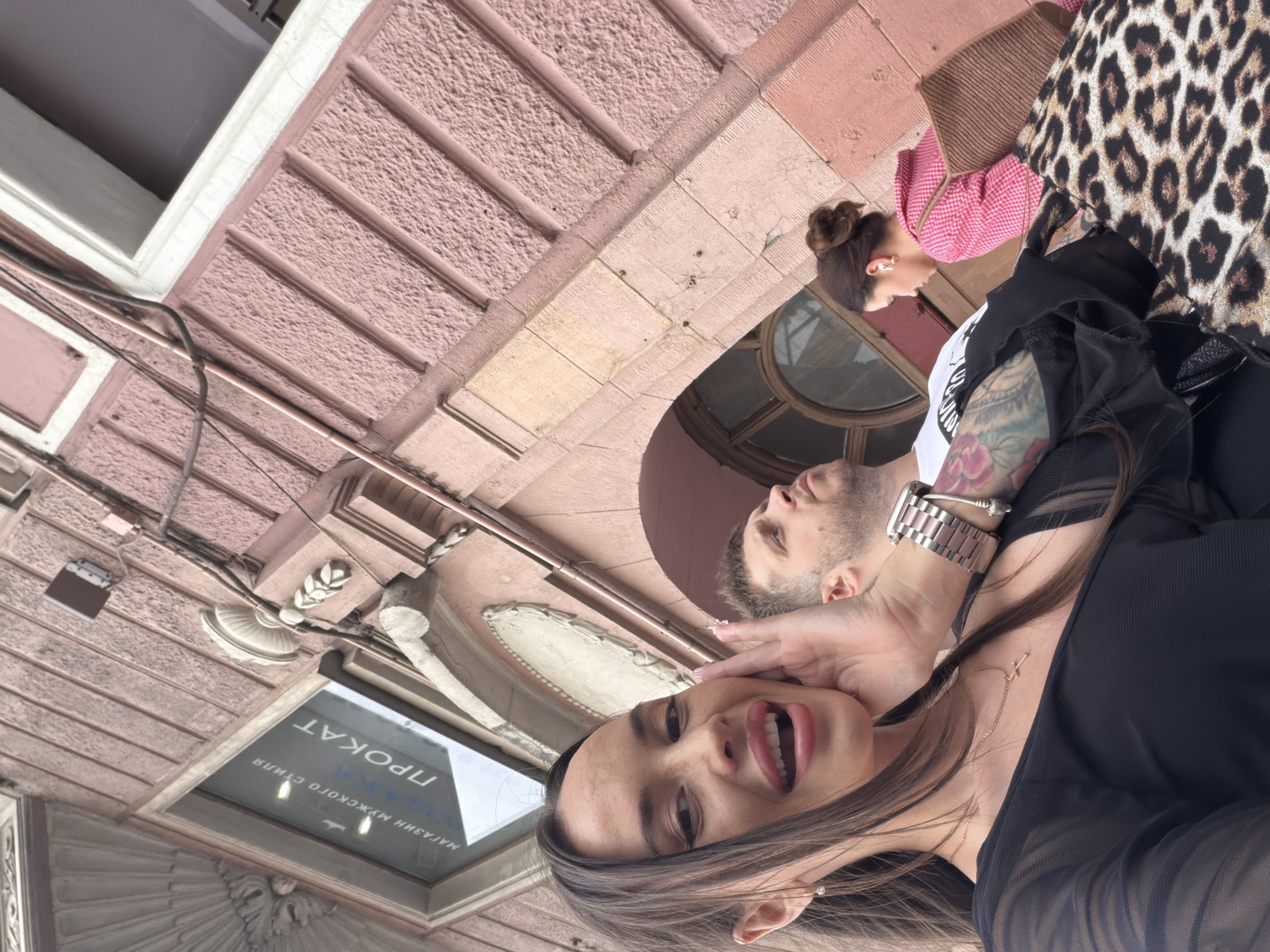 This street-level image captures three individuals in Saint Petersburg, Russia, set against a salmon-pink building facade. In the foreground, a woman with long dark hair, wearing a black sheer-sleeved top and a gold cross necklace, smiles widely with her mouth open, her right hand positioned near her cheek. She has a metallic watch on her left wrist and a prominent floral tattoo on her left forearm. A leopard-print bag rests in the lower right. Behind her, a man with short dark hair and a beard, wearing a white t-shirt and a dark-patterned tattoo on his left arm, looks upwards and to the right. Further back and to the right, a third woman with brown hair tied in a bun, wearing a pink checkered top and white wireless earbuds, carries a woven brown shoulder bag while looking downwards. The building features textured, vertically paneled stucco walls, white decorative cornices, and an arched window opening. Above the foreground figures, a shop window displays Cyrillic text, including "МАГАЗИН МУЖСКОГО СТИЛЯ" (MEN'S STYLE STORE) and "ПРОКАТ" (RENTAL), confirming the Russian context. Exposed pipes and wiring run along the building's exterior.