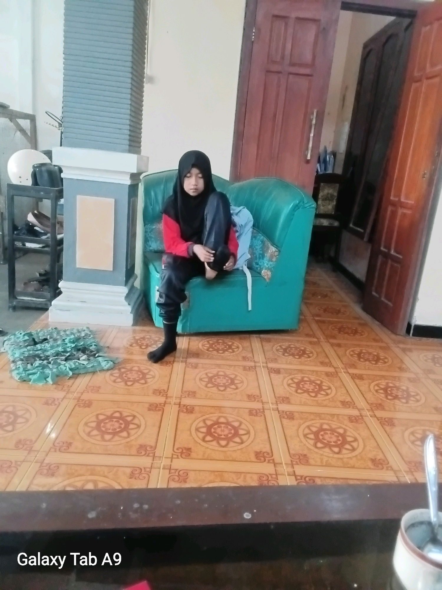 A young person wearing a black hijab, a red and black long-sleeved shirt, and black pants sits on a teal-colored sofa. The person is facing down with their hands in their lap. To the left of the sofa, a light grey pillar with textured detailing stands. In front of the pillar and to the left of the sofa is a small, green, woven mat. The floor is covered in light brown tiles with an ornate circular pattern. Behind the sofa, a brown wooden door is partially open, revealing a dark interior. The scene appears to be indoors, possibly in a residential setting. The image includes a timestamp or device identifier that reads "Galaxy Tab A9" in the bottom left corner. The specific location in Trenggalek, Indonesia, is not discernible from the visual information alone.