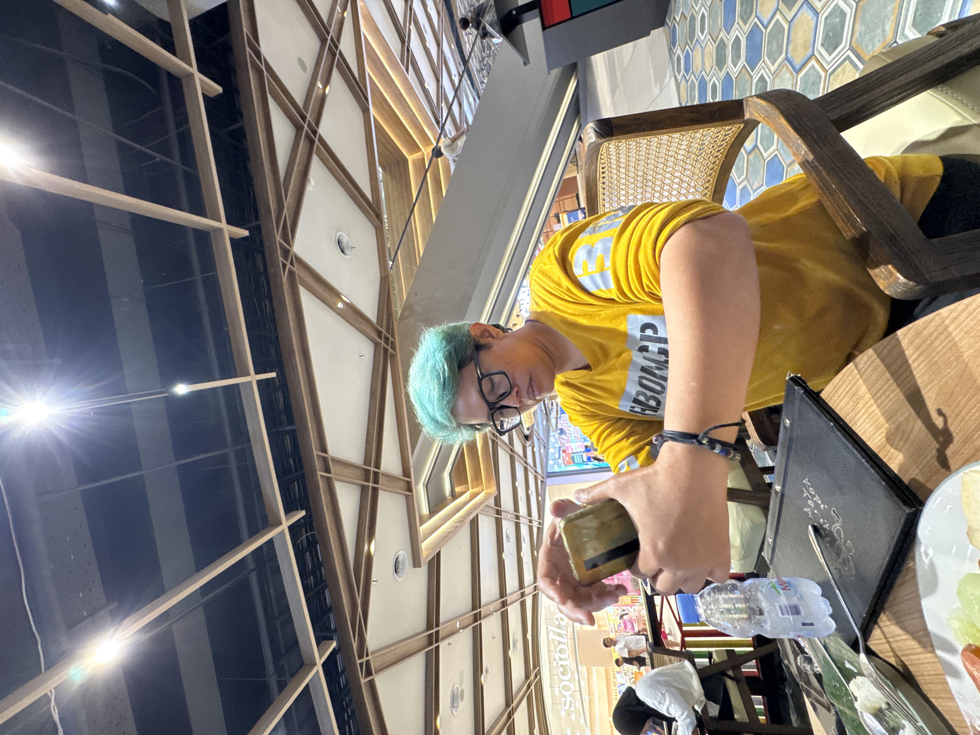 A person with turquoise-colored hair and glasses is sitting at a table in what appears to be a food court or restaurant within a shopping mall. They are wearing a yellow t-shirt with white lettering that reads "GBCNP". The person is holding a mobile phone in their hands, seemingly interacting with it. In front of them on the table is a menu, a bottle of water, and silverware. Other people are visible in the background, some dining and others walking. The ceiling features a complex wooden lattice structure with lighting fixtures. The floor has a hexagonal tile pattern in blue, green, and beige. The mall setting suggests this is likely a commercial area in Gunung Putri, Indonesia.