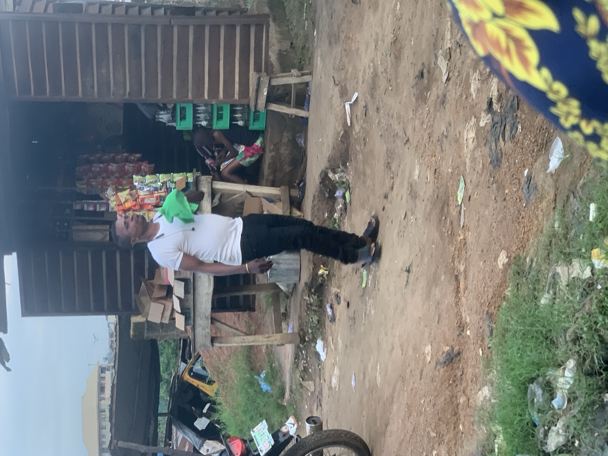 A man wearing a white t-shirt and black trousers stands in front of a shop. He holds a green cloth in his left hand. Behind him, a person sits in the doorway of the shop. The shop has shelves stocked with bottles and bags of goods. The ground in front of the shop is dirt and littered with trash. To the left of the shop, a motorcycle and a yellow vehicle are partially visible. In the background, a building with a light-colored facade and a dark roof is visible. The sky is overcast.
