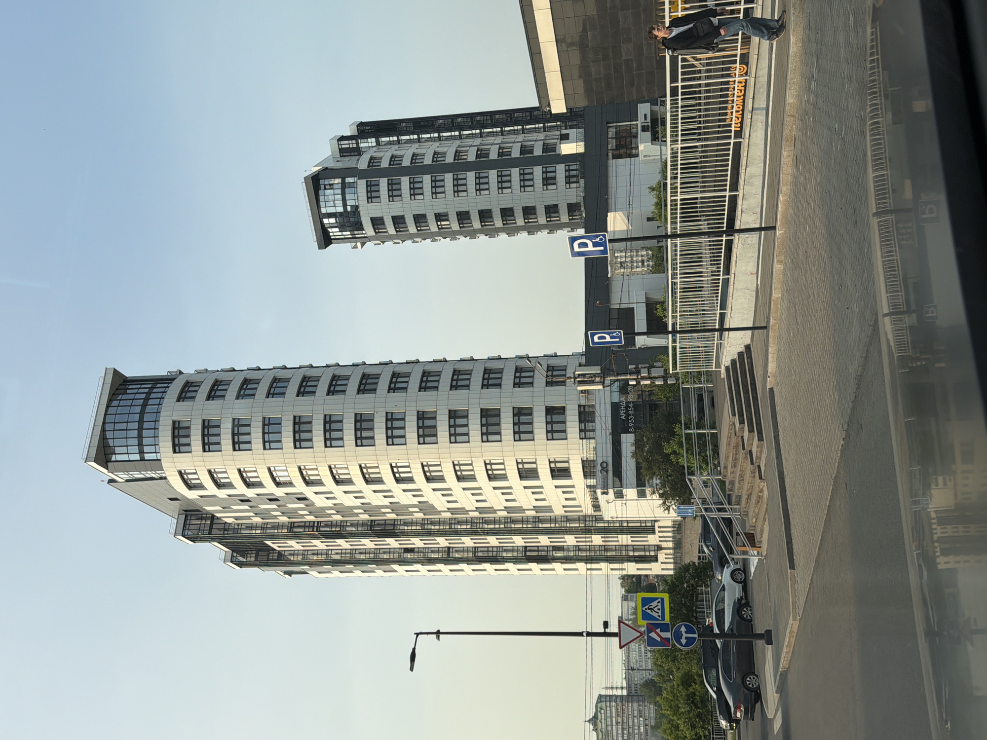 The image depicts a street scene in Krasnoyarsk, Russia, featuring two modern high-rise buildings. The larger building on the left is a curved, white, multi-story structure with numerous windows. The smaller building to its right is also a multi-story, modern design with dark and white paneling and glass elements. A street with cars, traffic signs, and a pedestrian walkway is visible in the foreground. A lone individual, appearing to be a young person, walks along the sidewalk on the right side of the frame. The sky is clear and pale, suggesting either early morning or late afternoon. The overall impression is one of urban development and daily life in a Russian city.