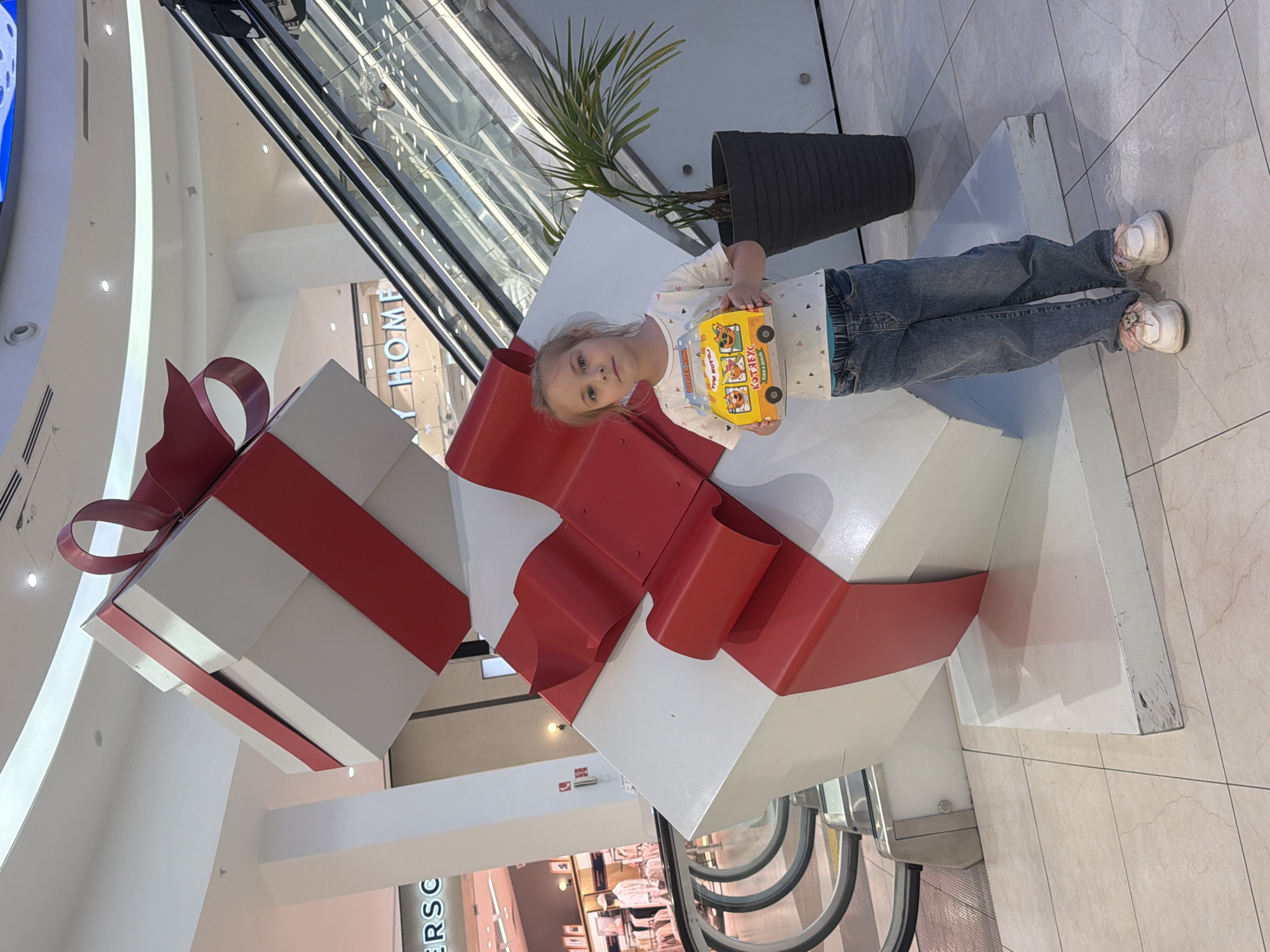 A young girl stands next to a large decorative stack of white gift boxes wrapped with wide red ribbons and bows, seemingly within a shopping mall. The girl is holding a yellow toy bus. She is wearing blue jeans, a white shirt with small hearts, and pink and white sneakers. The mall interior features an escalator behind the girl and the gift box display. A plant in a grey pot sits nearby. The tiled floor suggests a public space within the mall. The location is Novoaleksandrovka, Russia.