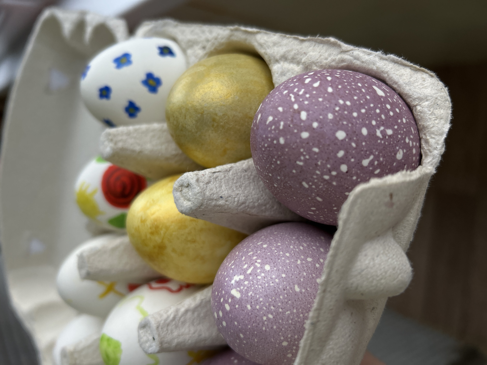 The image shows a close-up view of several decorated Easter eggs in a cardboard egg carton. The eggs are painted in different colors and patterns, including floral designs, a golden egg, and purple eggs with white speckles. The carton is held or displayed at an angle, showcasing the arrangement of the eggs. The location is likely not Chaikovsky, Russia as the context does not provide any details that would indicate this location.