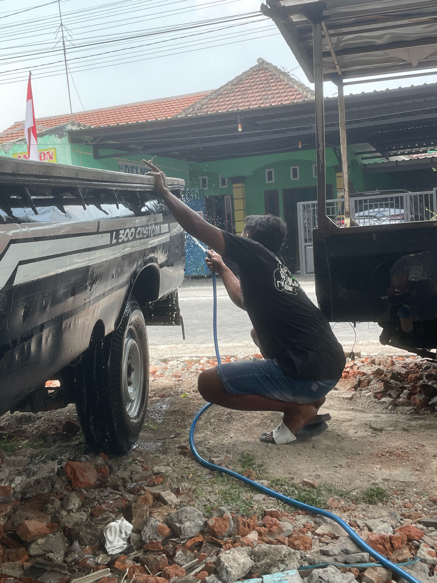 A man wearing a black t-shirt and blue shorts is crouched down and washing a black pickup truck with a blue hose in front of a building. The building is painted green and has a red tile roof. A red and white Indonesian flag is flying on a pole near the building. The ground is covered in dirt and rubble.