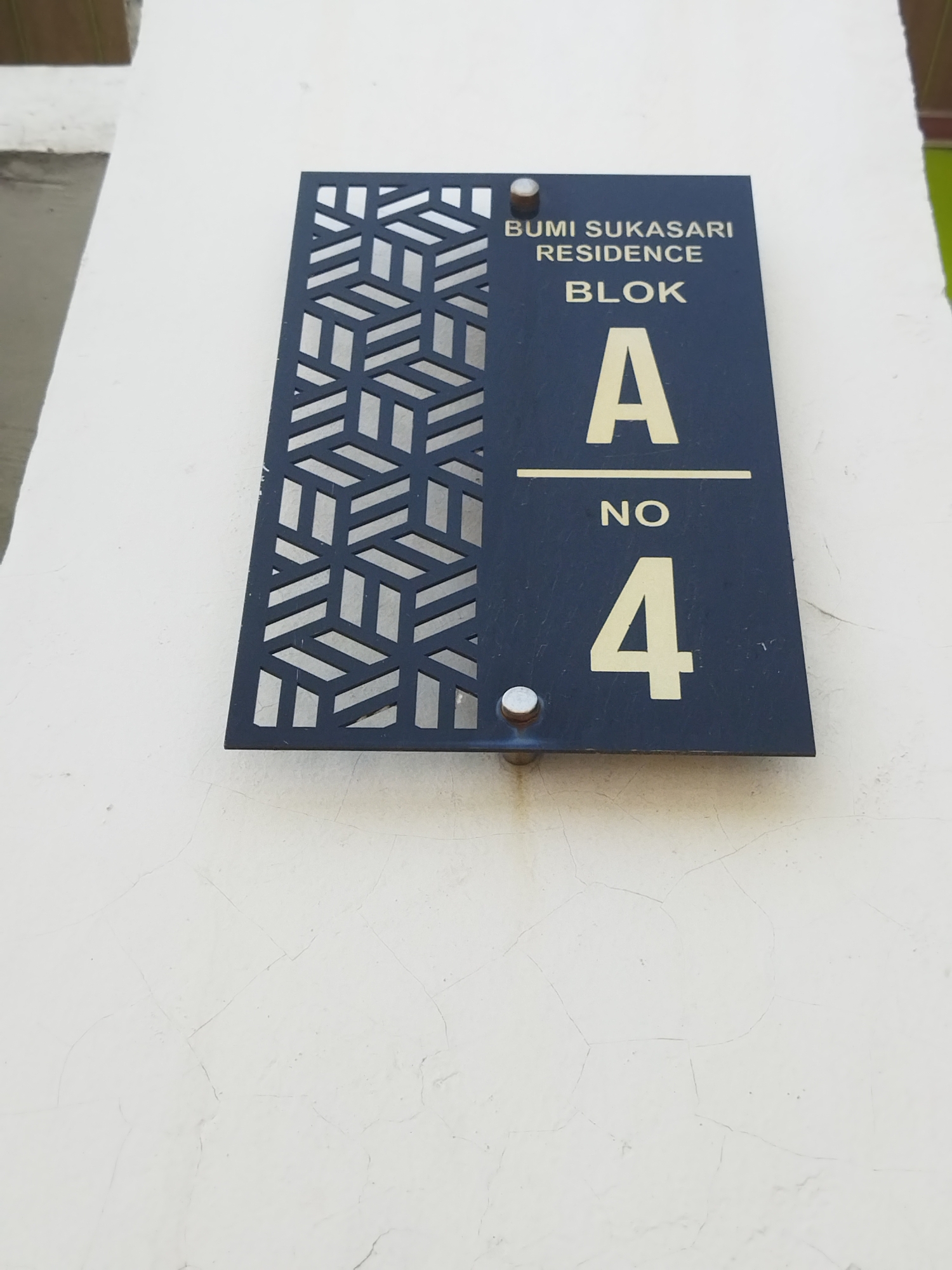 The image shows a close-up of a sign mounted on a white wall. The sign is dark blue and rectangular with a decorative geometric pattern on the left side. The right side of the sign displays text and numbers indicating an address. The text reads "BUMI SUKASARI RESIDENCE", followed by "BLOK A", and then "NO 4". The sign appears to be made of metal and is attached to the wall with two visible silver screws. The white wall has visible cracks and texture. The background is out of focus and shows a hint of a building's exterior with a shadowed area and a sliver of green. The image is taken from a slightly upward angle.