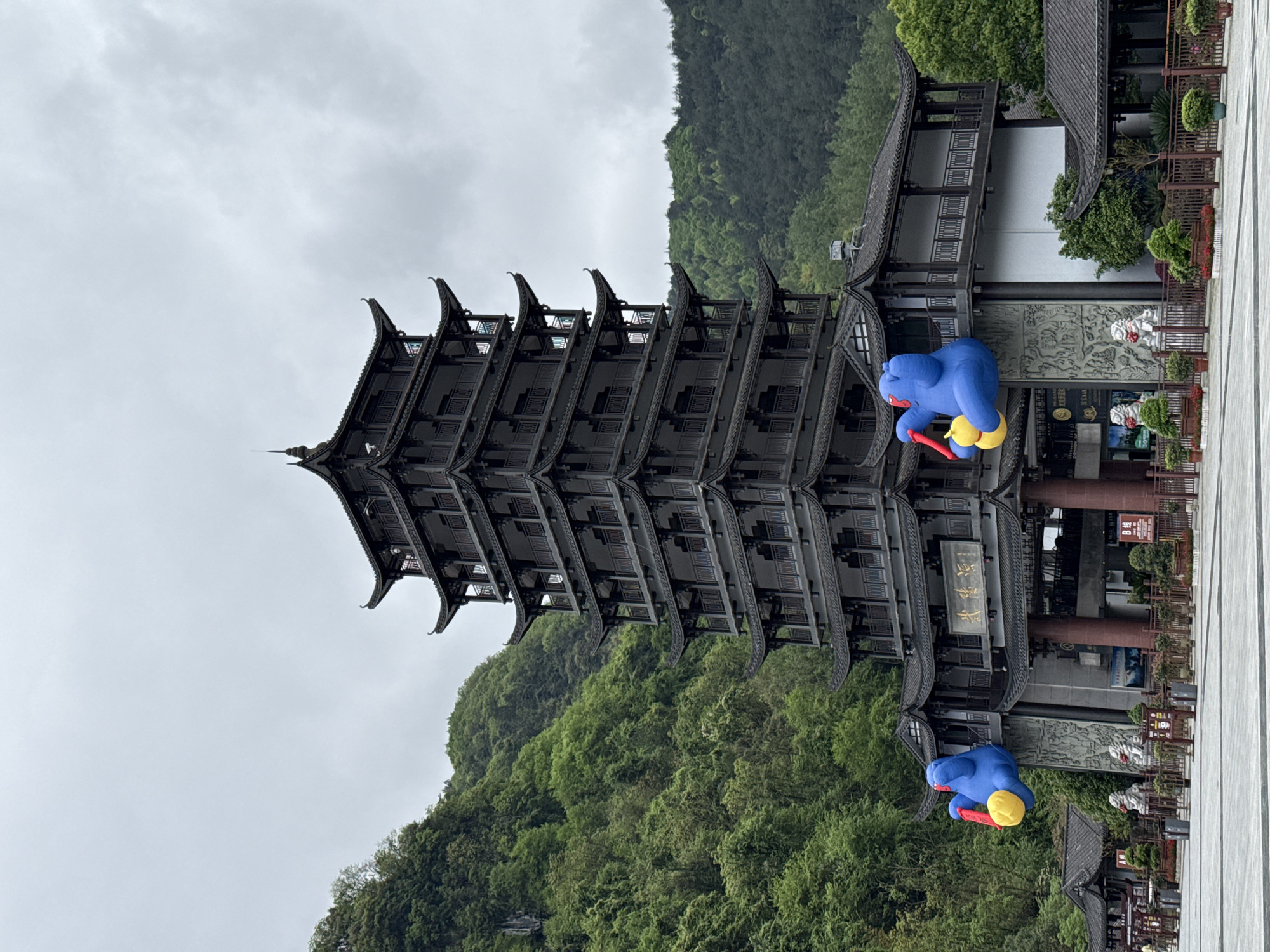 The image captures a multi-tiered traditional Chinese pagoda-style building set against a backdrop of a lush, green hillside and an overcast sky. The building has a dark, multi-layered roof with prominent upturned eaves. To the side of the pagoda is a lower building. Near the lower building are two large blue inflatable figures with yellow heads and red tools or accessories. The building is in the Wulingyuan Scenic Area, in Wulingyuan, China.