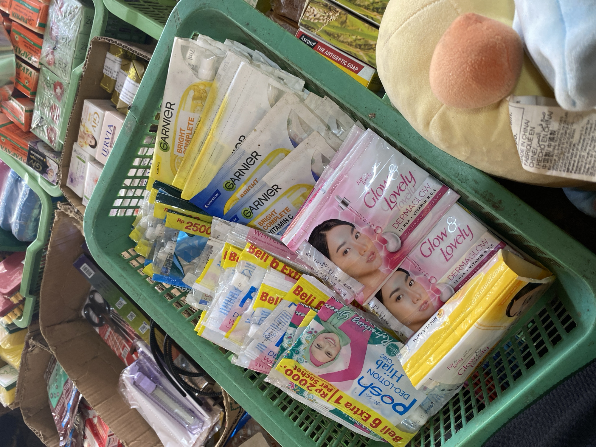 A close-up overhead shot shows a green plastic basket filled with various personal care products, likely in a market stall in Ta', Indonesia. The basket contains numerous small sachets and packets of skincare items, including Garnier Bright Complete sachets and Glow & Lovely Dermaglow sachets. Also visible are Posh Deo-Lotion Hijab Chic sachets. In the background, other baskets and boxes hold more items, such as cleaning brushes and packaged soaps. A yellow plush toy with an orange circular nose is partially visible in the lower right corner. The lighting is bright, suggesting an indoor or shaded outdoor location.