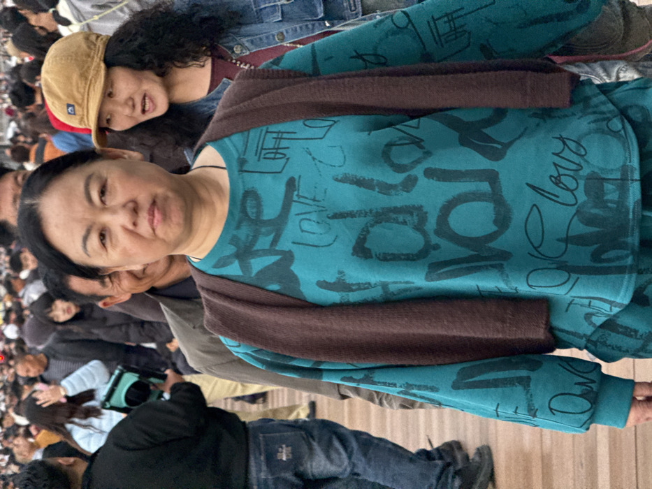 The image displays a densely populated public area within Yuzhong District, China. Centered in the foreground is a middle-aged East Asian woman looking directly forward with a neutral expression. She is wearing a teal long-sleeved top adorned with black graffiti-style text, including the word "LOVE," and a dark brown sleeveless garment draped over her shoulders. To her immediate right, a partial view of an East Asian man's face and shoulder is visible, with a slight smile evident. To her left, a younger East Asian woman with dark, curly hair and a tan baseball cap is partially observed, her mouth slightly open as if in conversation, dressed in a denim jacket over a dark red top. The background is filled with a large crowd of East Asian individuals, all casually dressed, suggesting a busy thoroughfare. On the far left, an individual is seen holding a teal-colored electronic tablet. The ground surface is composed of light-colored wooden planks. The scene is illuminated by ambient light.