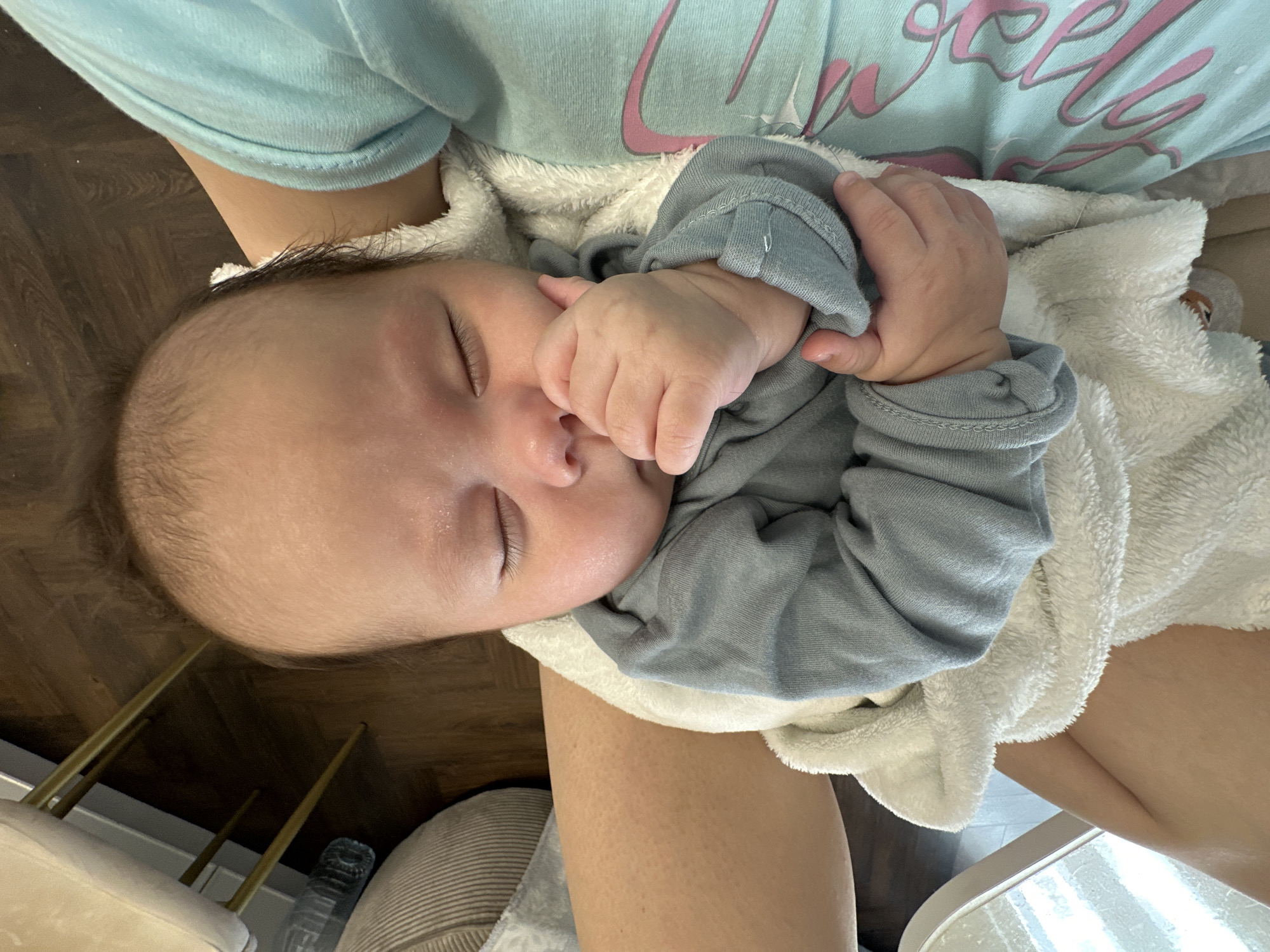 This close-up image captures a peaceful moment of an infant sleeping soundly while being held by an adult. The baby, appearing to be a newborn or young infant with fair skin and fine dark hair, has closed eyes, indicating deep slumber. One tiny hand is curled up near its face, adding to the sense of comfort. The baby is dressed in a light gray long-sleeved outfit and is partially covered by a soft, white, fuzzy blanket or wrap.
The adult holding the baby is partially visible, with a light blue long-sleeved shirt or sweatshirt, featuring some indistinct light pink cursive text. Parts of the adult's arm and leg are also seen, suggesting the baby is cradled or resting on their lap. The setting appears to be indoors, with dark wooden flooring visible in the background. Other elements in the periphery include slim, golden-colored metallic rods, possibly furniture legs, a partially visible clear water bottle, and a segment of light-colored, ribbed fabric from what might be an armchair or cushion. The overall emotion conveyed is one of tranquility and tenderness, focusing on the infant's serene repose. No specific time of day or location is discernible beyond an indoor domestic setting.