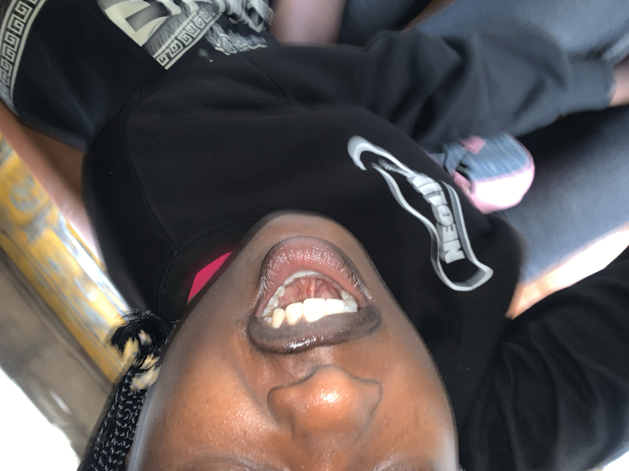 A close-up, tilted, first-person perspective shot shows a Black individual with their mouth open, revealing teeth. The individual's head is angled downwards, and their face is in focus from the nose down. They are wearing a black long-sleeved shirt with a white graphic and a pink inner layer. A small pink and blue bag is visible to the lower right of the frame. The background is blurred and appears to be a yellow-tinted surface, possibly a vehicle interior, with hints of brown and grey. The image appears to be taken indoors or in a sheltered environment. There is no clear indication of the specific location within Jos, Nigeria, beyond the inferred context of the prompt.