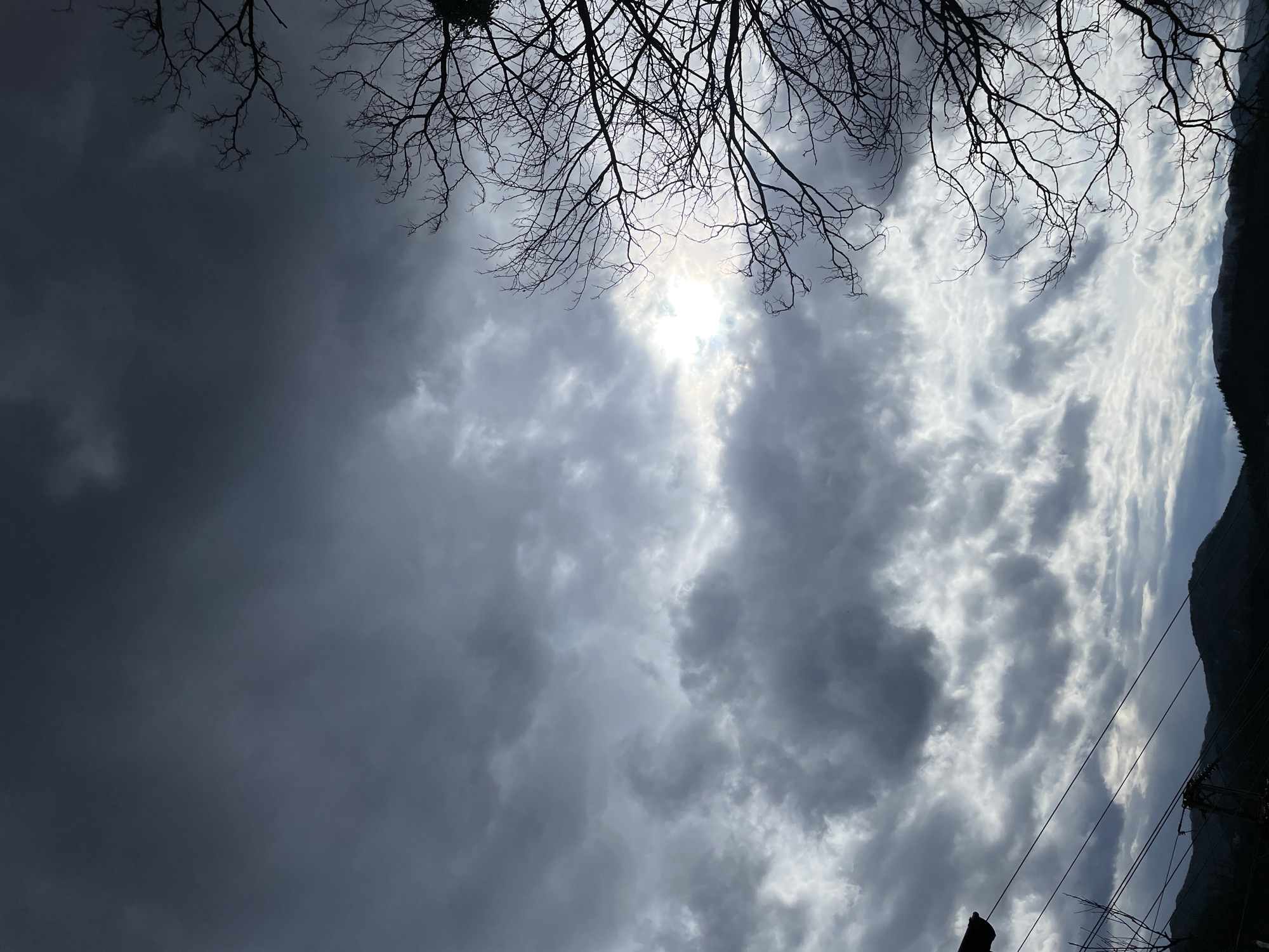 The image depicts a sky filled with dark, grey clouds. The sun is partially obscured by the clouds, creating a diffused light. Silhouetted tree branches are visible in the upper portion of the image. A dark, mountainous shape is present on the right side of the image with powerlines are in the foreground. The location is likely Zlatograd, Bulgaria given the landscape and weather.
