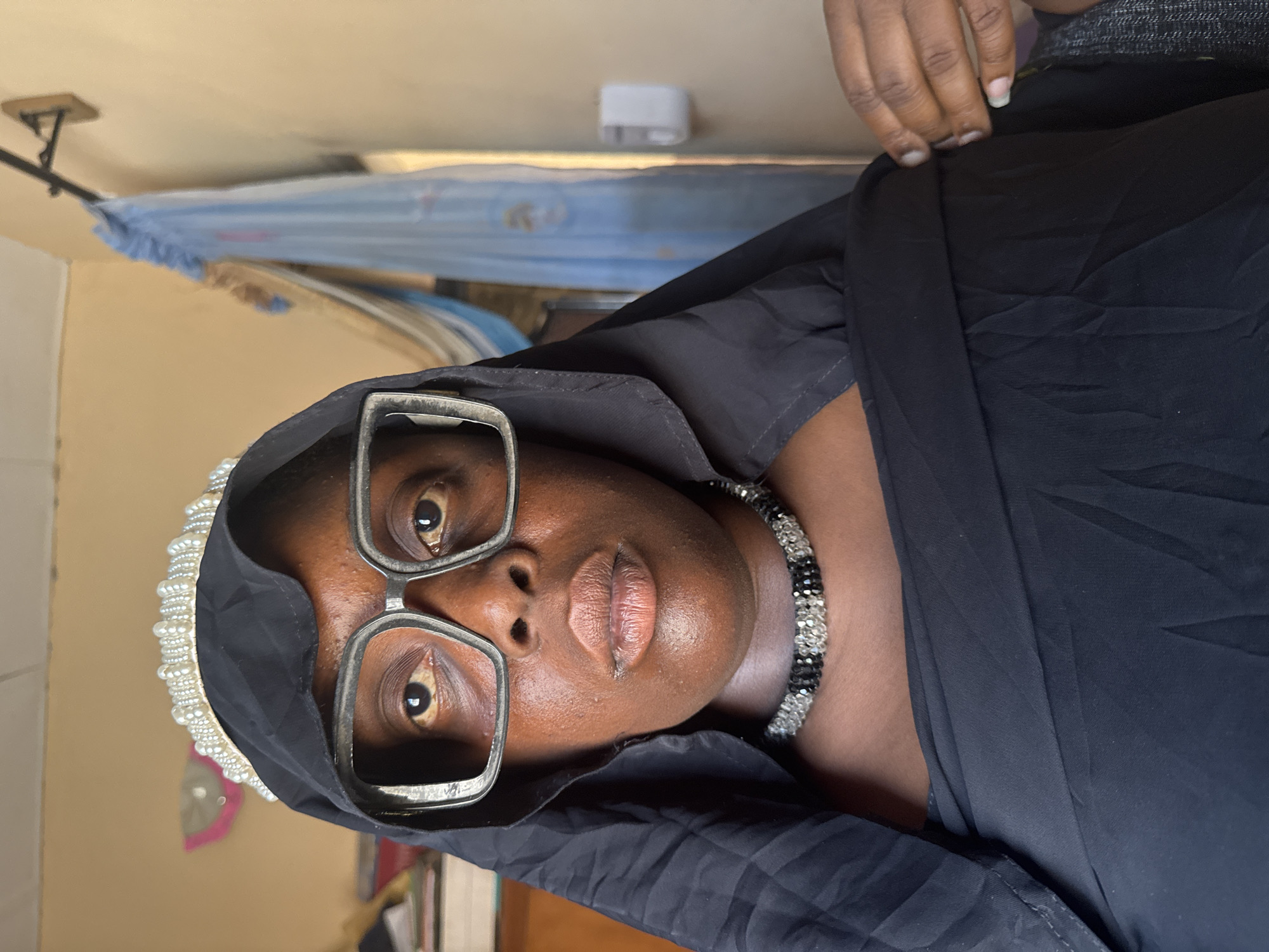 A medium shot shows a dark-skinned individual wearing large, silver-framed glasses and a black head covering. A string of black and white beads is visible around the individual's neck. Part of a pearl-studded headpiece is visible to the left of the head covering. The individual is indoors with what appears to be a mosquito net hanging in the background. Other objects in the background include a small white device and what appear to be books or papers. The scene might be inside a home or room in Madangala, Nigeria. There is no evidence to confirm this.