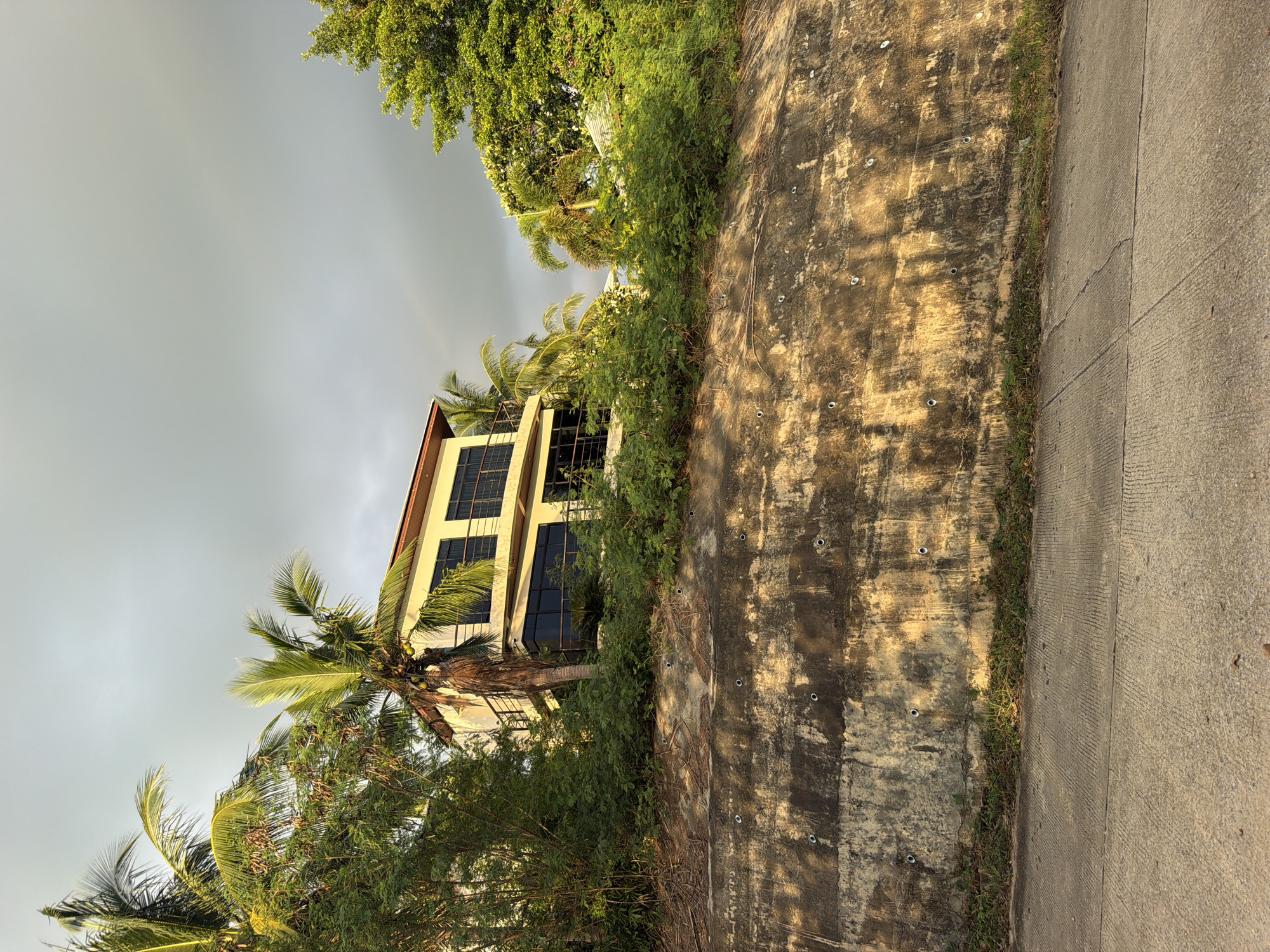 The image depicts a modern, two-story house situated on a hillside surrounded by lush tropical vegetation, including palm trees and dense foliage. The house features large, dark-tinted windows and balconies. In the foreground, a concrete retaining wall slopes upwards, covered in patches of dry grass. Beyond the wall, a paved road is visible. The sky is overcast with some lighter patches, and a faint rainbow is visible in the distance. The lighting suggests it is either early morning or late afternoon, with golden sunlight casting shadows. The scene appears to be a residential area in Baan Thong Po, Thailand, characterized by its natural surroundings and contemporary architecture. No people or animals are present in the frame.
