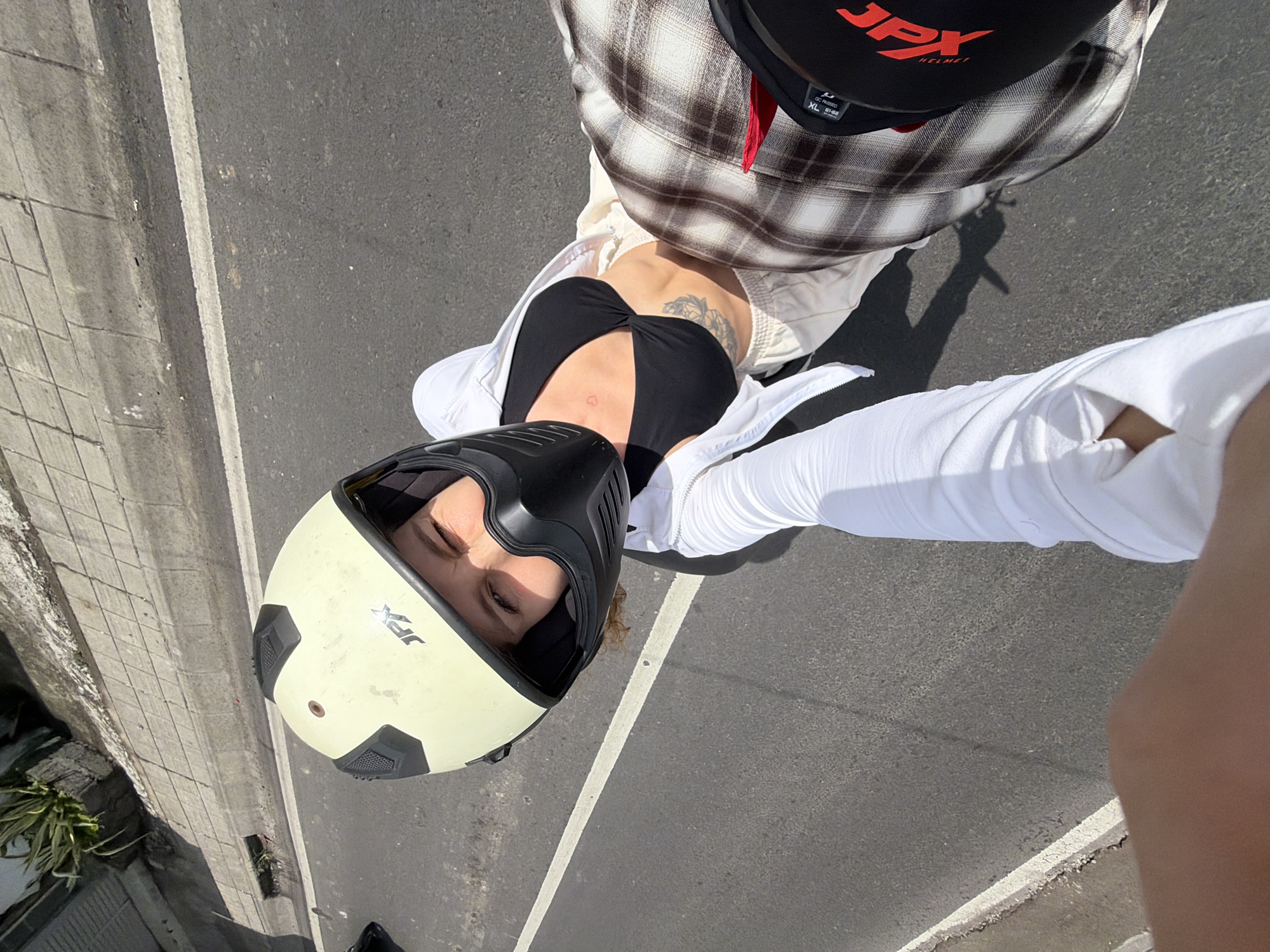 The image shows two people on a motorcycle in North Kuta, Indonesia, on a paved road with a white line marking a lane. The person in the foreground is wearing a white long-sleeved top and has their arm extended. The person in the background is wearing a light-colored helmet with a black face shield and a black bikini top. They are also wearing light-colored pants and a plaid shirt. A tattoo is visible on the person in the background's torso, and a smaller heart-shaped tattoo is on their chest. In the upper left corner of the image, a planter with a plant is visible next to a concrete sidewalk. The lighting suggests it is daytime.