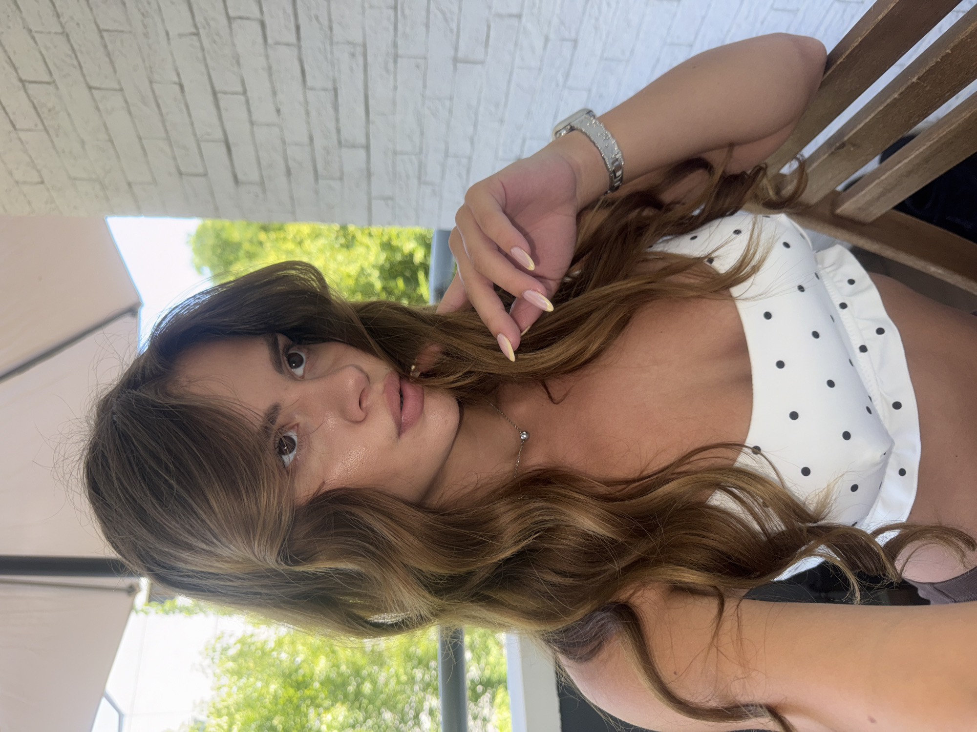 A young woman with long, wavy brown hair is shown in a close-up, upper-body shot. She is looking to her right, with her left hand resting near her face. She is wearing a white polka-dot top and a silver necklace with a small pendant. A wristwatch is visible on her left wrist. The background includes a white brick wall, lush green foliage, and a portion of a large umbrella. The setting appears to be an outdoor seating area, possibly a balcony or patio. The image was taken on a sunny day, casting soft light on the woman's skin.
