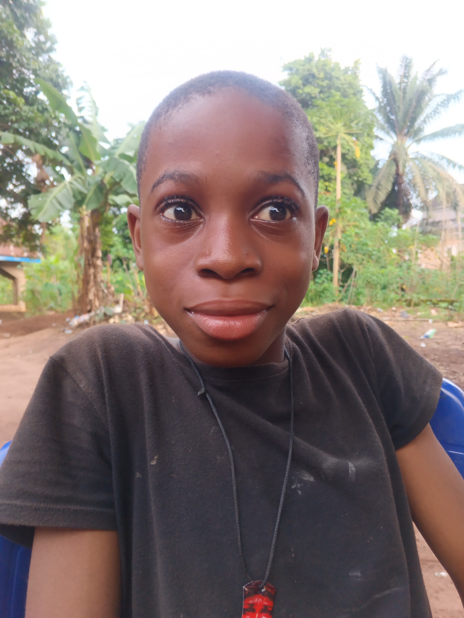A close-up portrait shot features a young person, likely a boy, with dark skin and short dark hair, looking subtly to the right with a faint smirk. His dark eyes also gaze to the side, conveying a playful or slightly shy expression. He wears a plain black t-shirt, which shows some light dust or marks, and a black string necklace with a distinctive red and black rectangular pendant. A portion of a blue plastic chair is visible behind his right arm and shoulder, suggesting he is seated.
The background is a vibrant outdoor scene, characteristic of a tropical or semi-tropical environment, consistent with Eziudo, Nigeria. Lush green foliage dominates, with broad banana leaves and what appears to be a palm tree prominently visible, indicating a rural or semi-rural setting. The ground is unpaved dirt. The lighting is bright and even, suggesting daytime conditions without direct harsh sunlight, possibly under an overcast sky or during the golden hour. The subject's skin has a natural sheen. There is no visible text in the image.