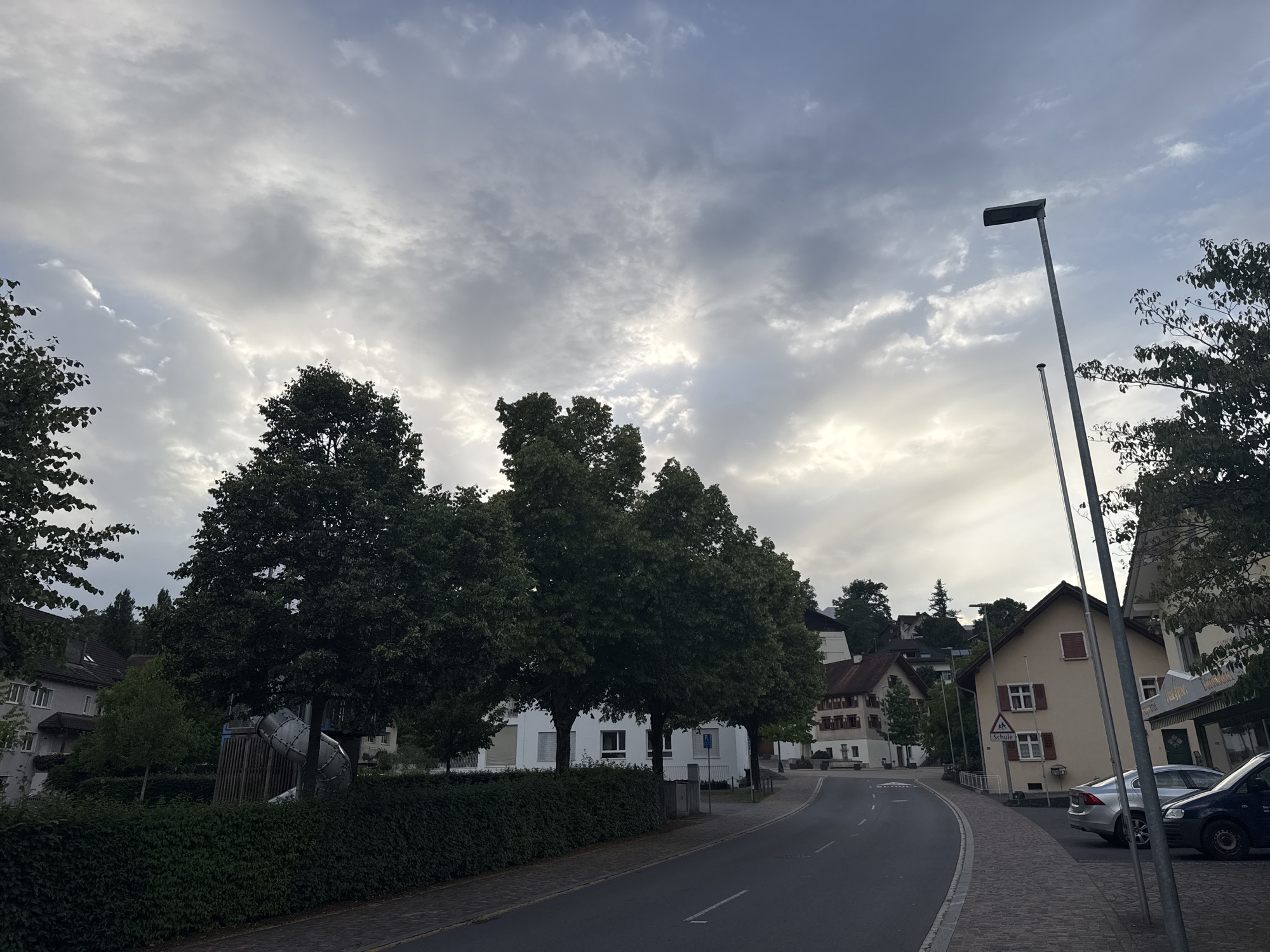 The image depicts a street scene in Nendeln, Liechtenstein, under a cloudy sky. On the left, a row of lush green trees lines a hedge. Behind the hedge, a playground structure with a silver slide is partially visible. Further back, a white building with windows is situated. To the right of the road, several houses are visible. The closest house is a light yellow building with red shutters and windows. A street sign with a school symbol is posted nearby. In front of the houses, a silver car and a dark blue van are parked. The road curves to the right, leading towards more houses and trees on a hill. A street lamp stands on the right side of the road.
