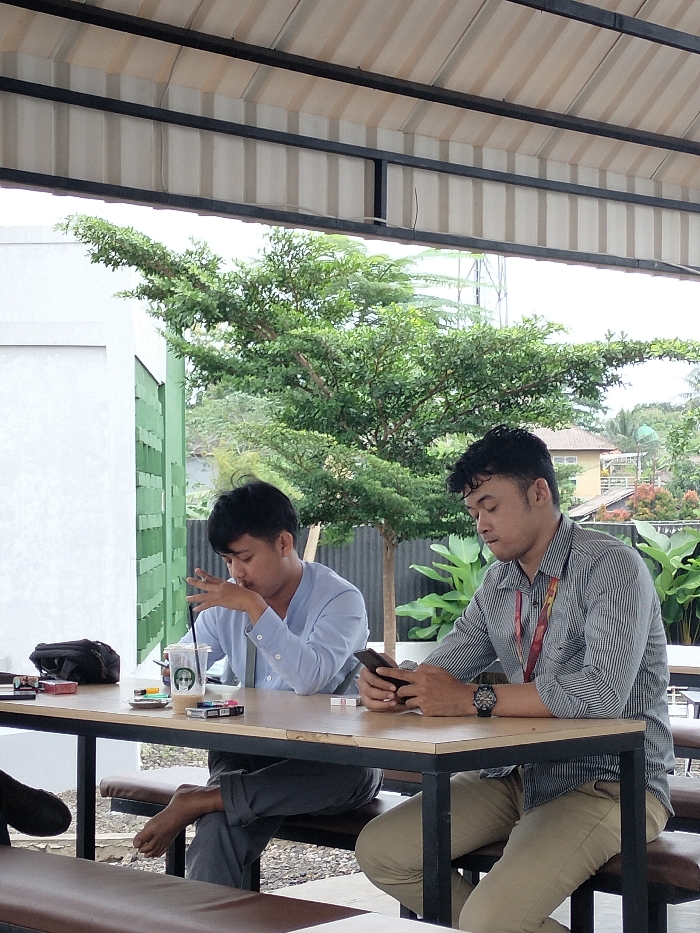 This image captures two young men seated at an outdoor or semi-outdoor cafe in Dramaga, Indonesia. The setting features a corrugated metal roof supported by black beams overhead, and lush greenery including a large tree dominates the background. The sky appears overcast, suggesting a mild, possibly warm day.
On the left, a man with dark hair, wearing a light blue shirt and grey pants, sits with one bare foot resting on the bench. He holds a clear plastic cup with a straw, adorned with a logo resembling Starbucks, and appears to be in thought or sipping his drink. On the wooden table in front of him, several packs of cigarettes are visible, including "Gudang Garam International" and another red and white pack, along with lighters and an ashtray. A black bag or pouch is also on the table.
To the right, another man with dark, curly hair, dressed in a striped shirt and khaki pants, is engrossed in his smartphone. He wears a watch on his left wrist and an ID badge on a red lanyard around his neck. Both men are seated at a wooden table with black metal legs, on benches with dark cushioning.
The background shows parts of a white and green building structure, and further back, glimpses of residential houses amidst more foliage. The ground beneath the table appears to be covered with pebbles or gravel. The overall scene suggests a casual break or a gathering, with both individuals engaged in their own activities.