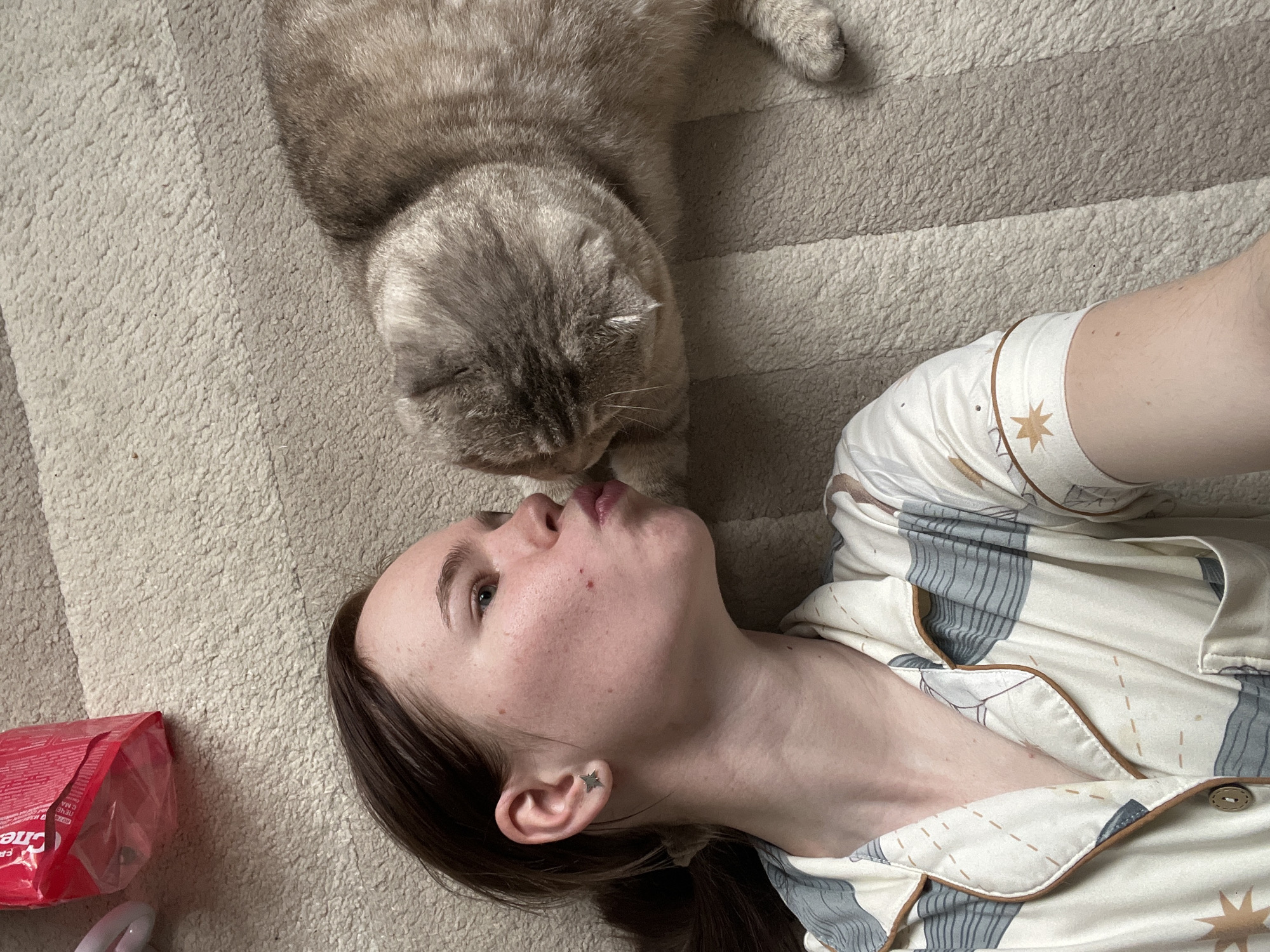 A woman is lying on a beige carpet, facing upwards. A gray tabby cat is positioned directly above her head, with its face near her lips. The woman is wearing patterned pajamas, beige in color with blue and brown accents. To the left of the woman’s head is a red package, partially visible. The carpet is a light beige color, with subtle striped patterns. The exact location in Chaikovsky, Russia cannot be determined from the image.